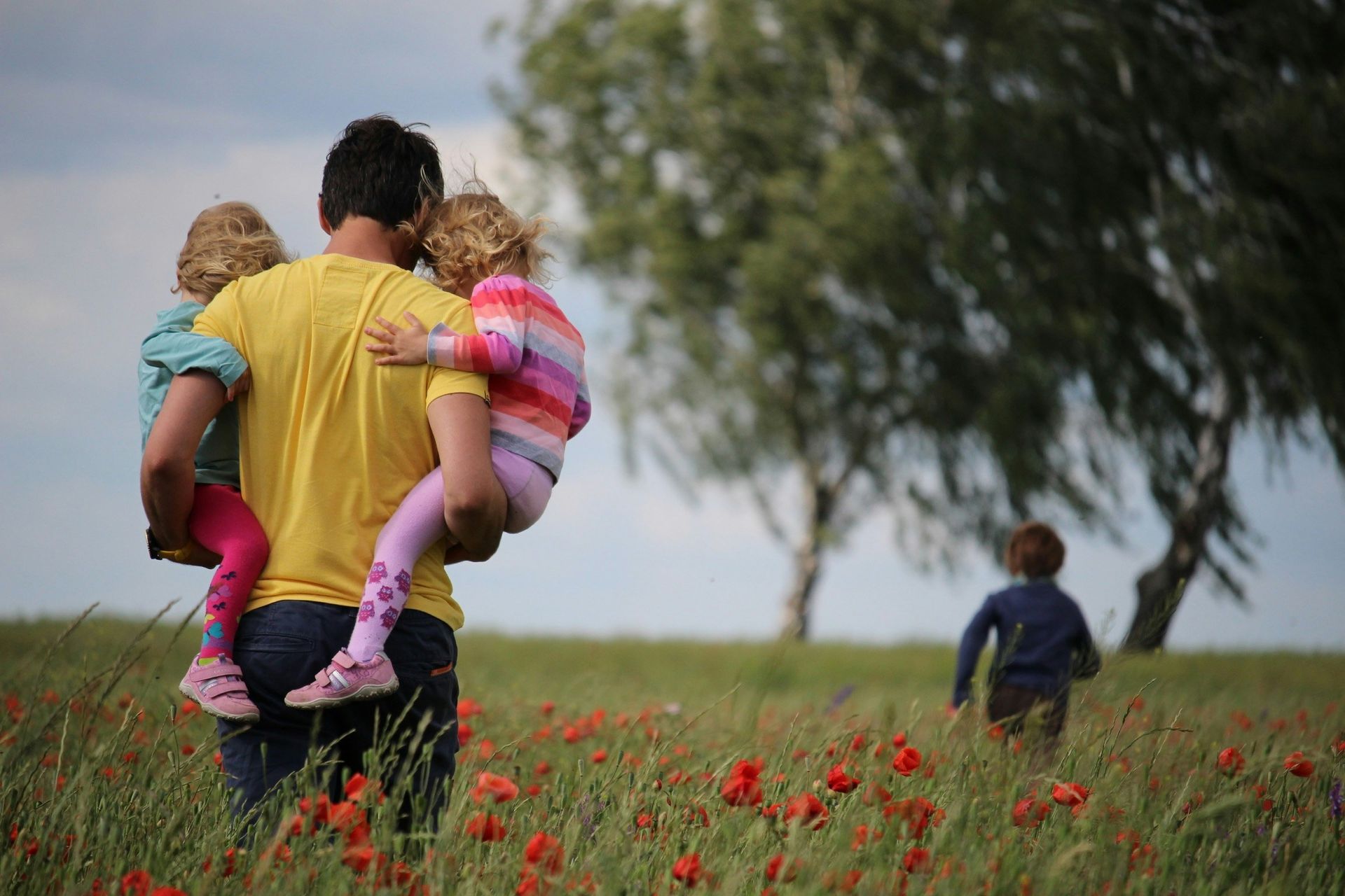 Man carrying two children in a field of red flowers; another child runs ahead.