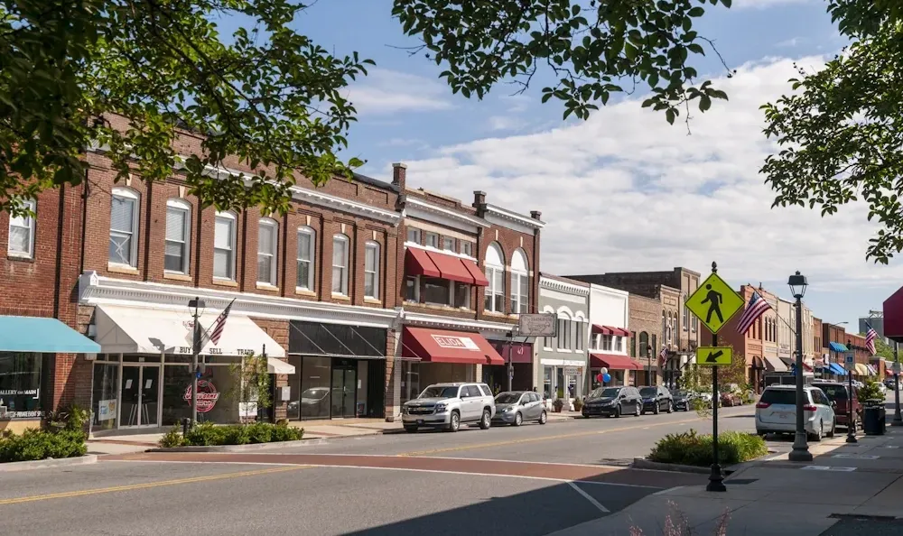 Street view of downtown buildings with shops, awnings, and parked cars on a sunny day.