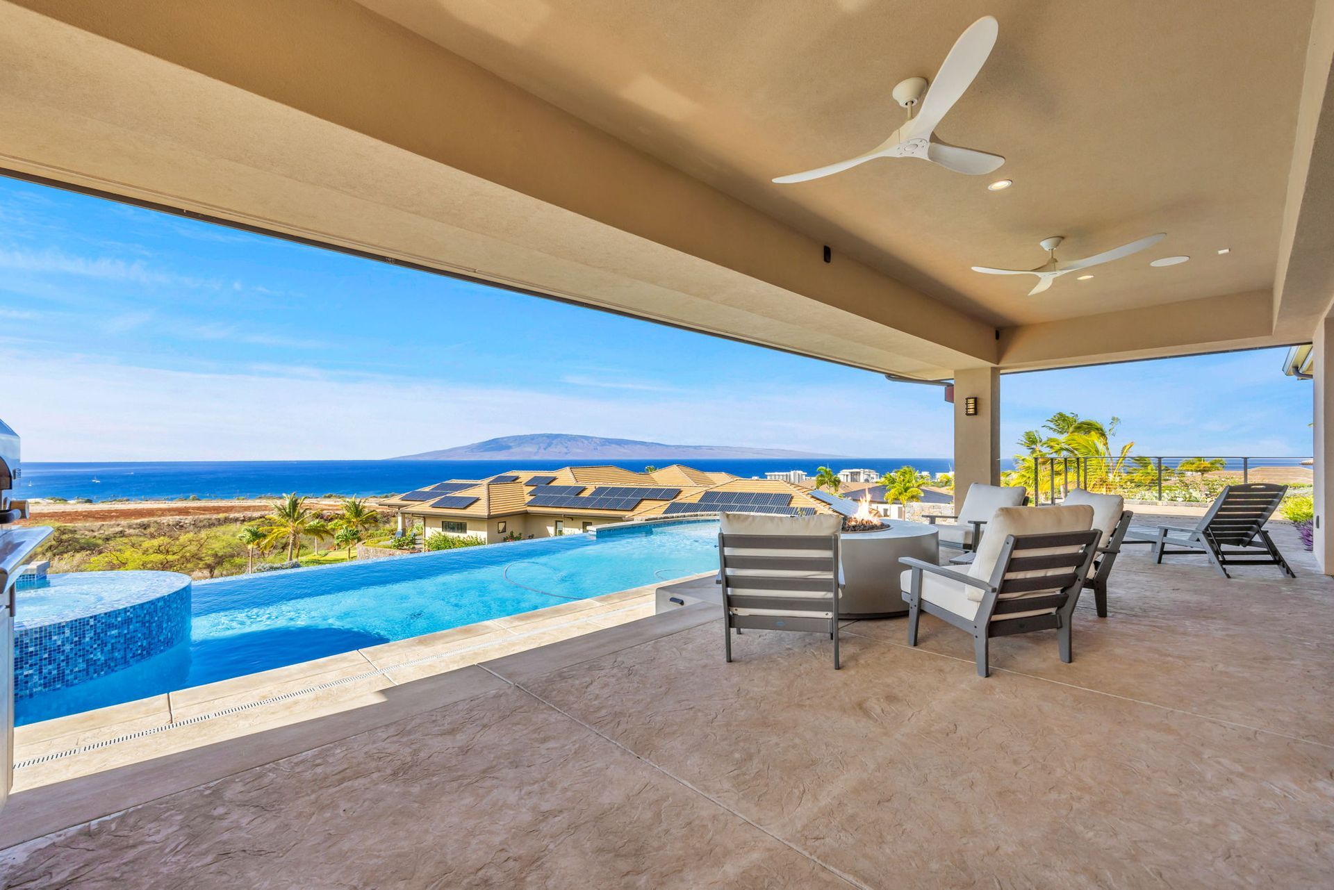 An open-air patio overlooking an infinity pool and the ocean, featuring outdoor seating under a covered ceiling.
