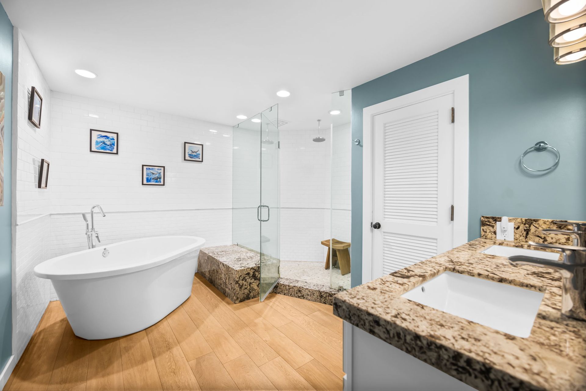 A modern bathroom featuring a white soaking tub, a stone-tiled walk-in shower, granite vanity, and light wood flooring.