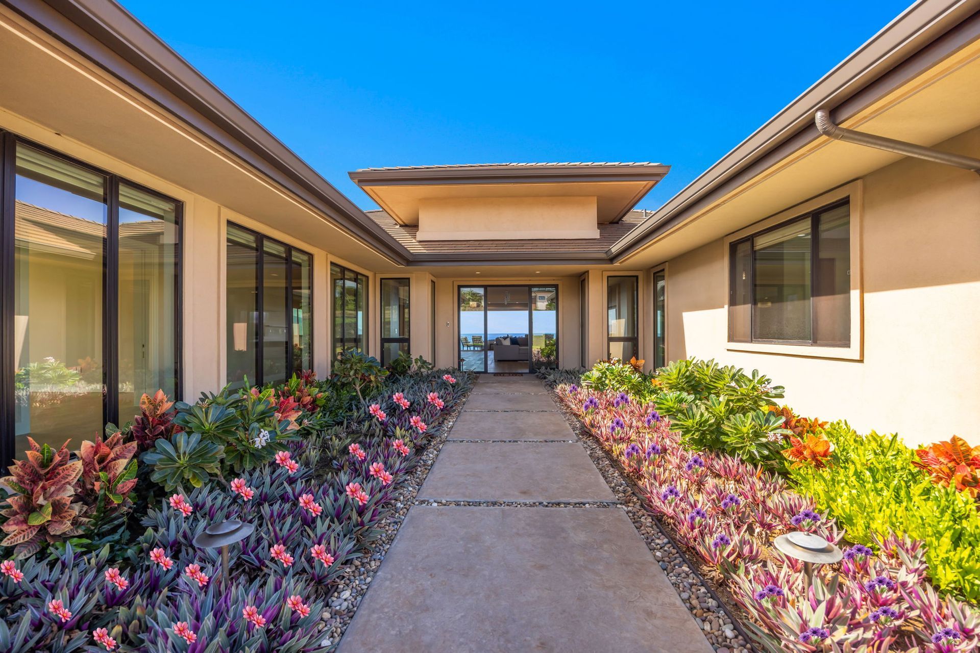 A stone pathway leads to the front entrance of a modern beige house, flanked by vibrant purple and green tropical plants.