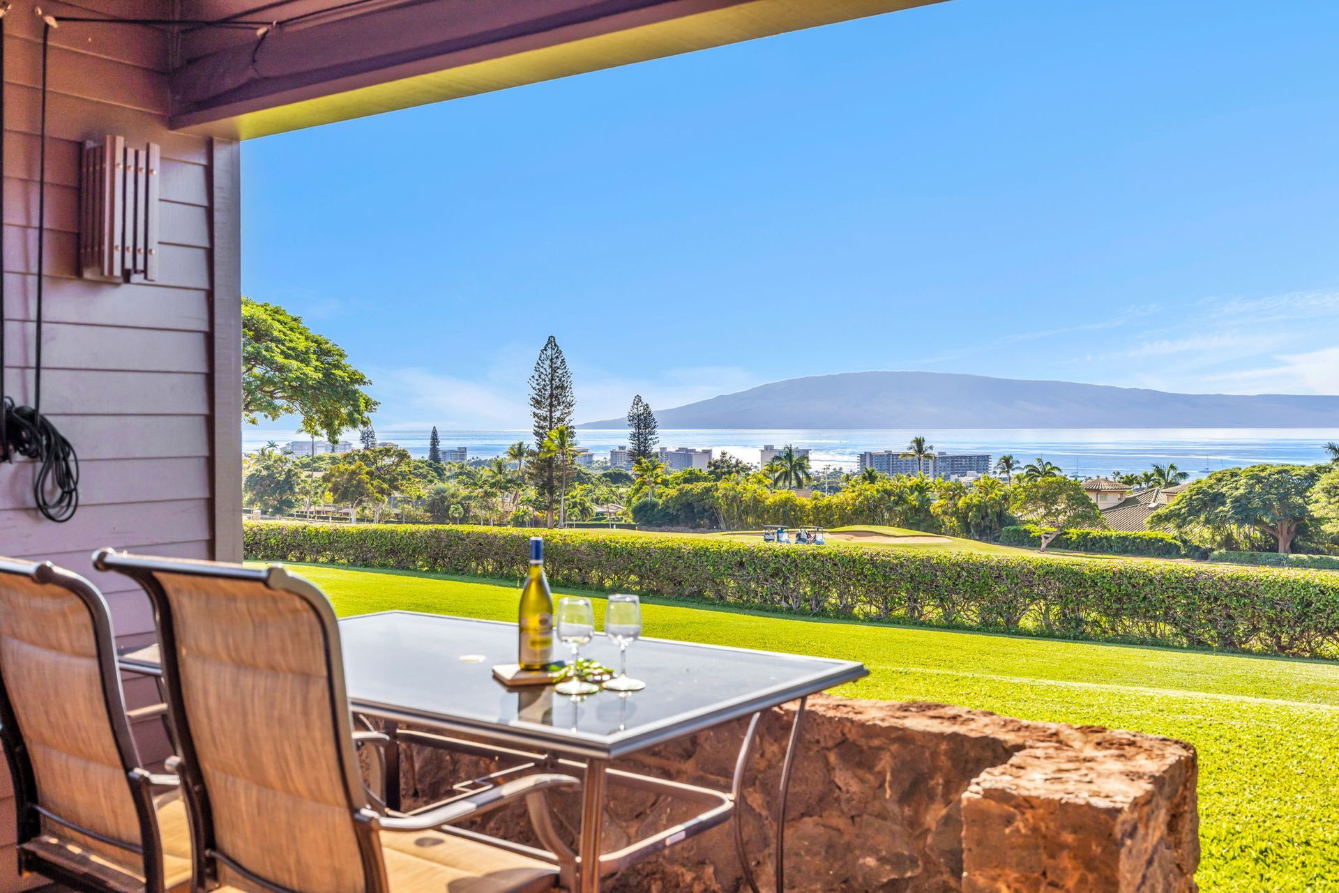 A glass patio table with wine and glasses overlooks a grassy lawn, palm trees, and an ocean view with mountains beyond.