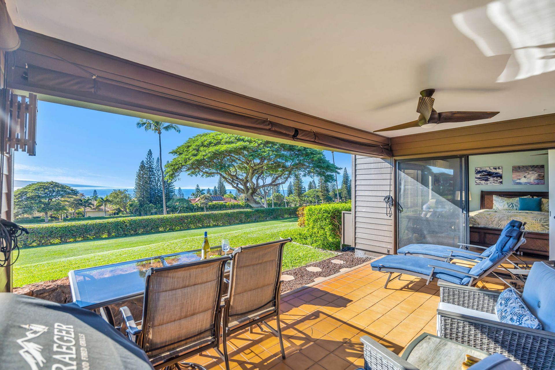 A covered outdoor patio with dining furniture and lounge chairs overlooks a lush grassy lawn and distant ocean view.
