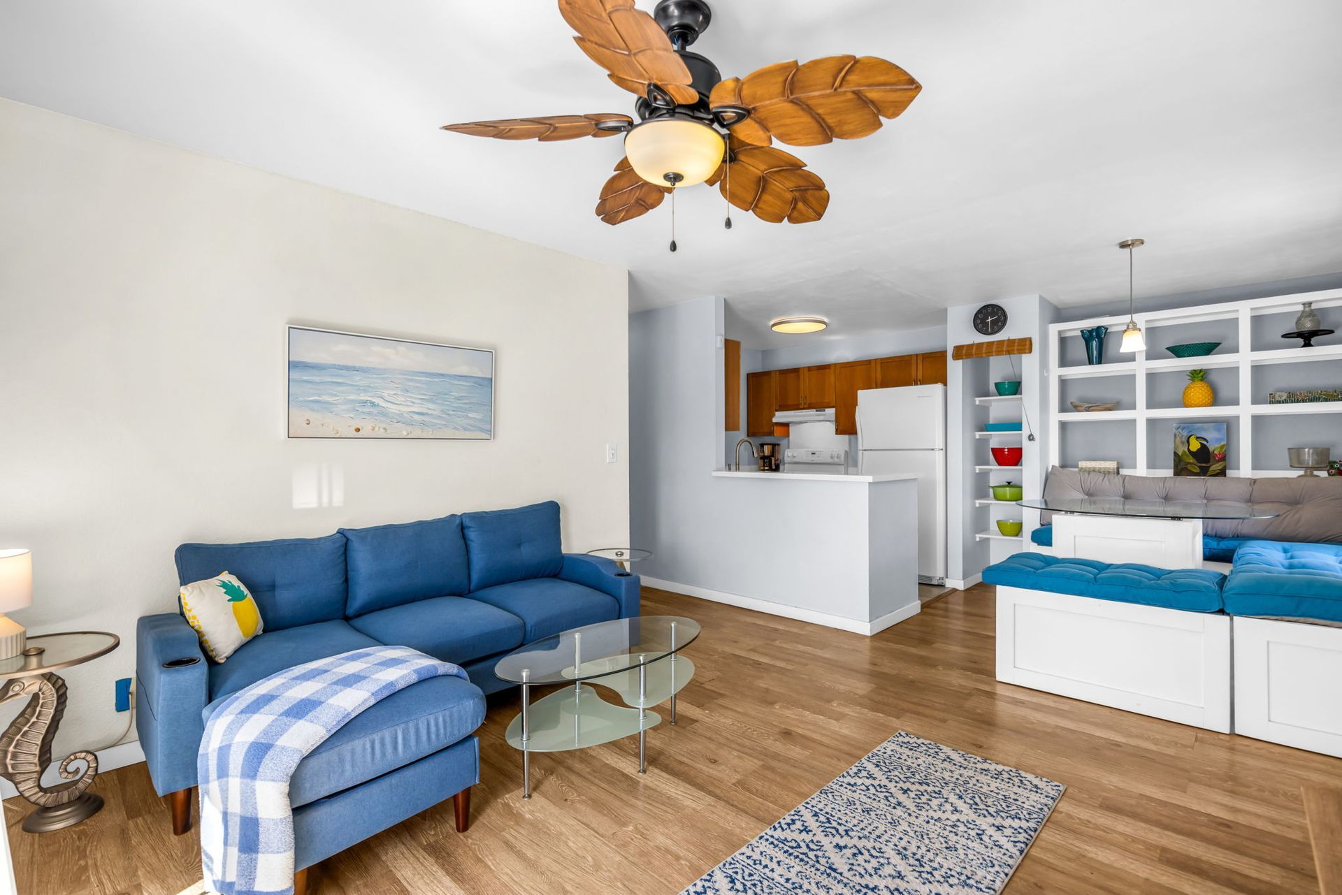 A living room with a blue sofa and ottoman, a glass coffee table, and a wooden ceiling fan, opening to a small kitchen.