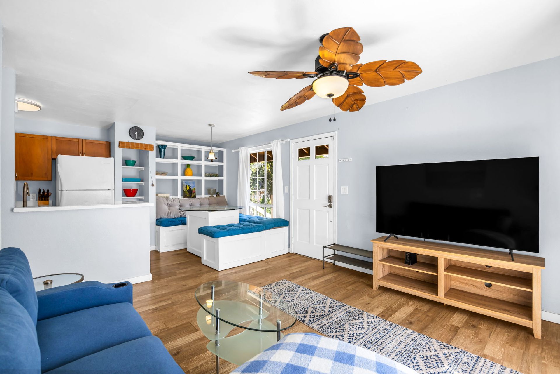 Living room with a blue sofa, large flat-screen TV on a wooden stand, and a dining nook with blue cushions.