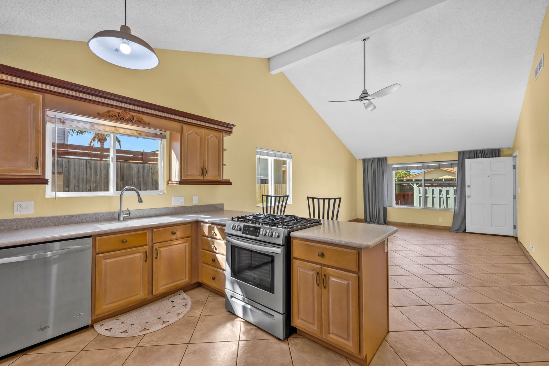 A bright kitchen with light wooden cabinets, stainless steel appliances, and beige tile floors under a vaulted ceiling.