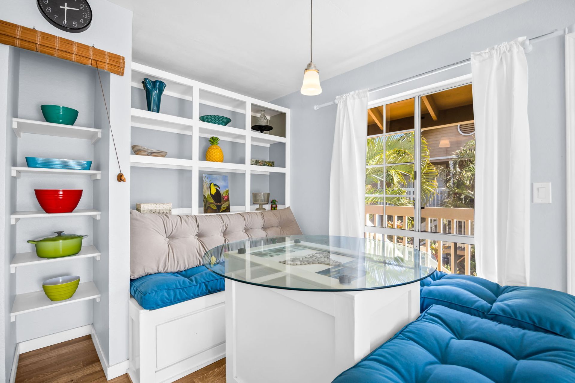 A breakfast nook with a glass table, blue tufted cushions, built-in shelving with colorful bowls, and a view outdoors.