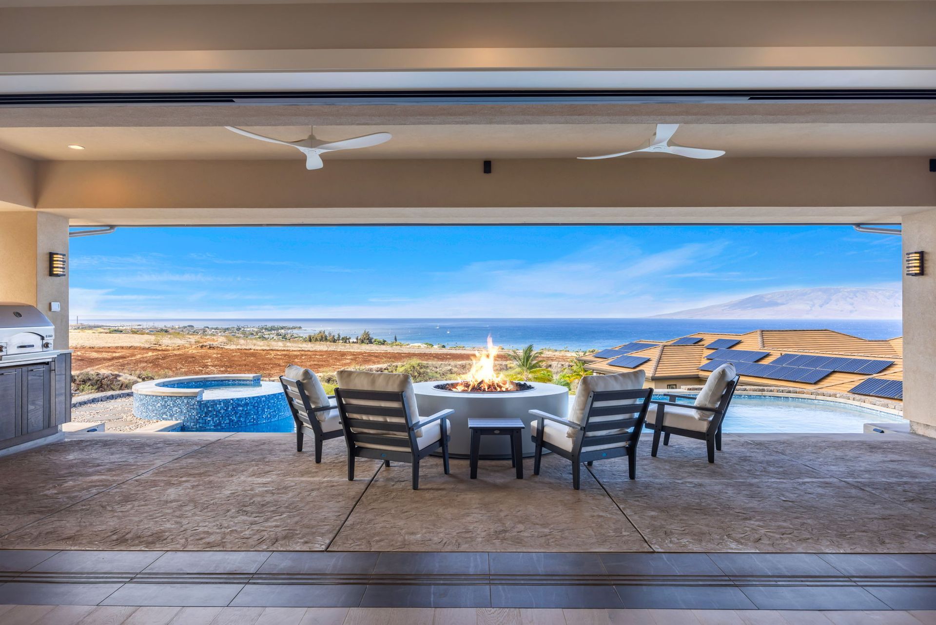 An outdoor patio with a fire pit and armchairs overlooks a swimming pool and the ocean under a clear blue sky.