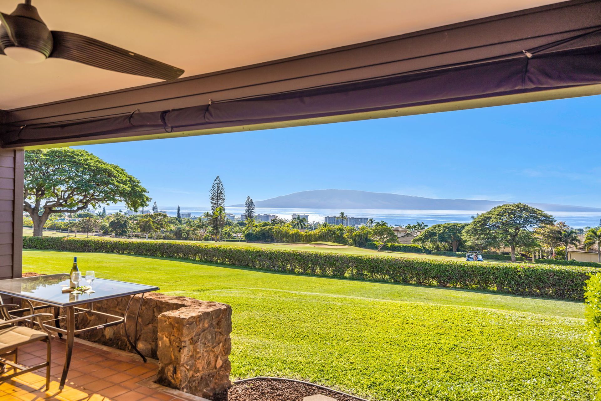 Patio with a dining set overlooking a sprawling green lawn, trees, and a distant ocean view under a clear blue sky.