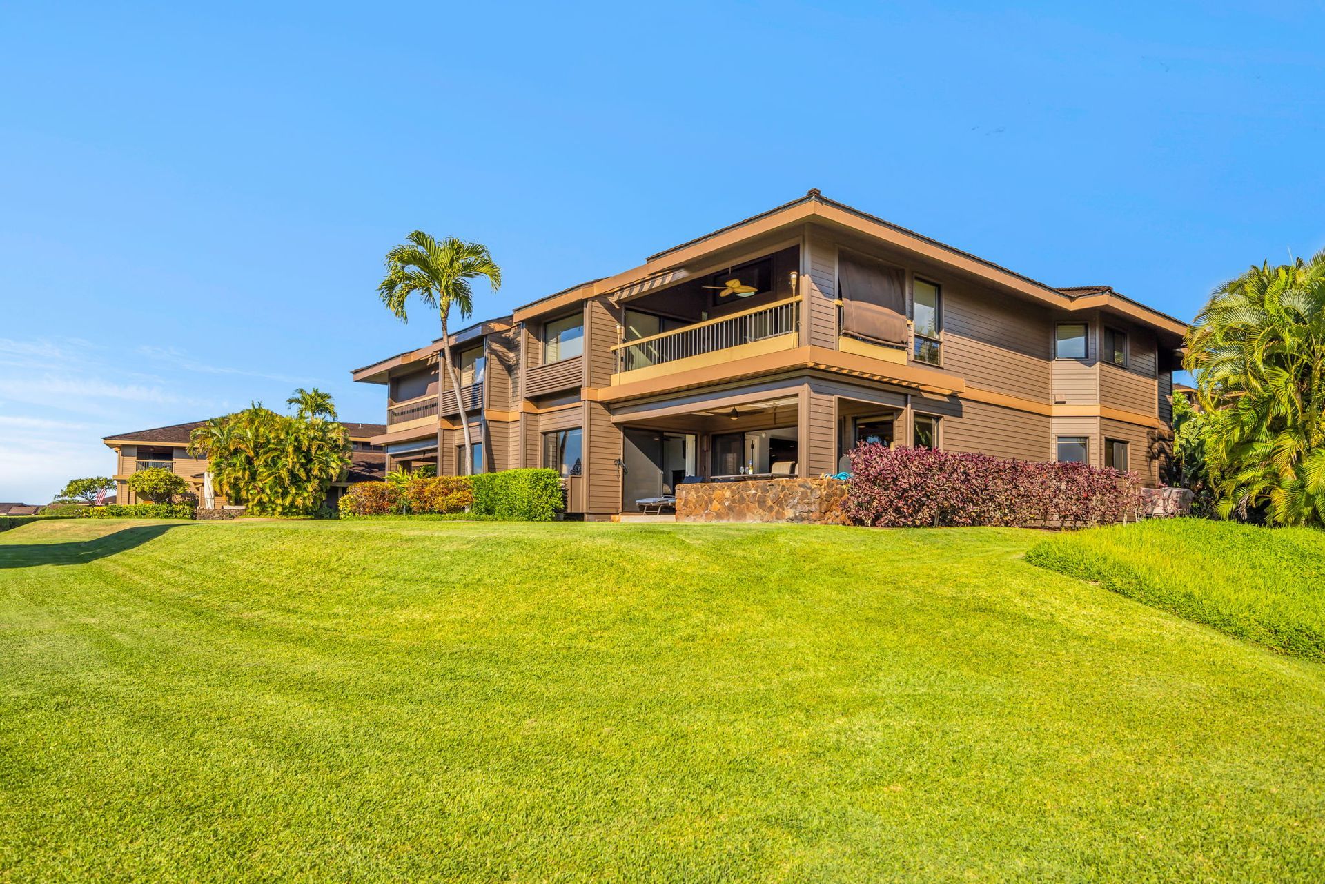 A two-story brown wood building sits behind a wide, lush green lawn under a clear blue sky, surrounded by palm trees.