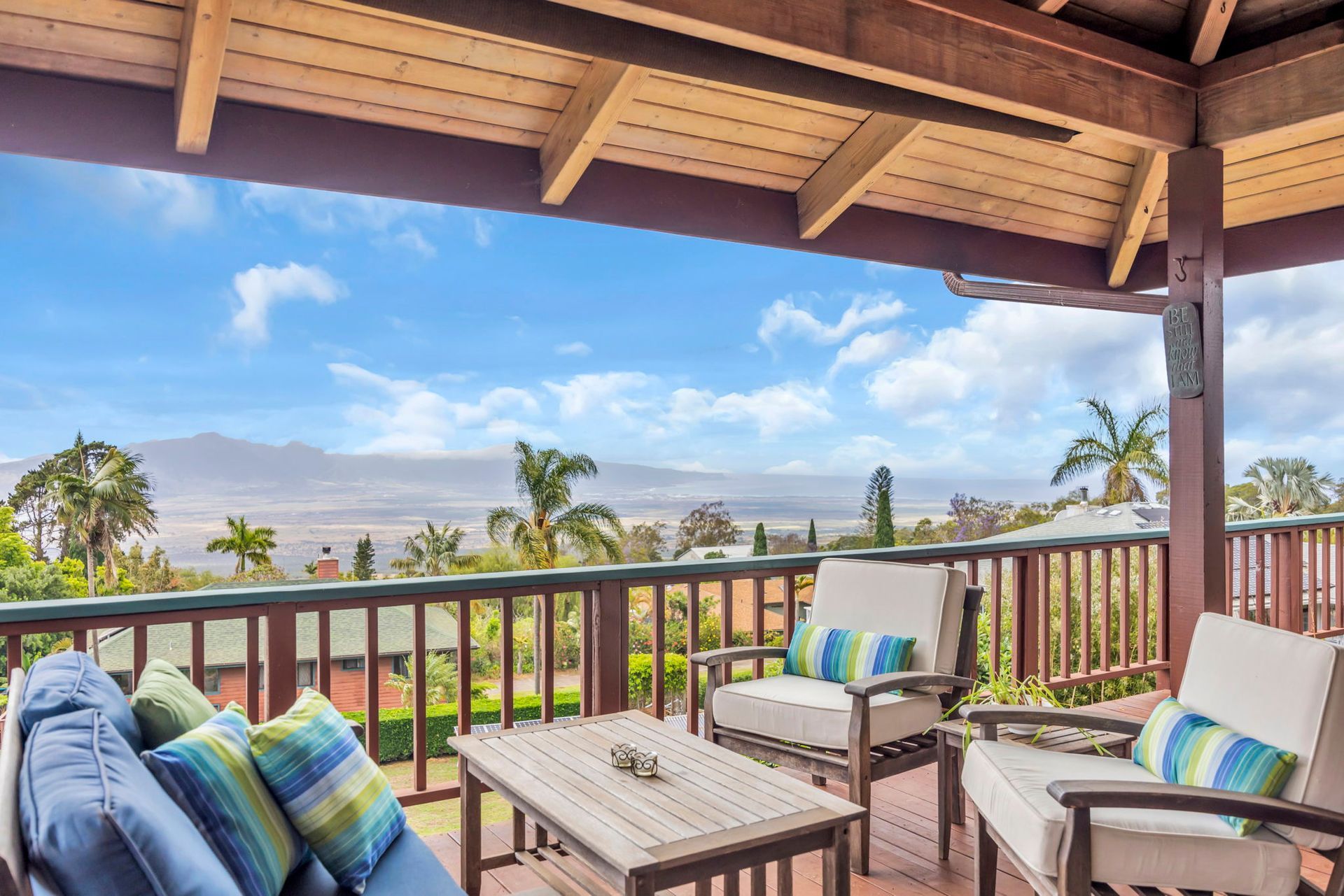 A covered wooden deck features two armchairs and a blue sofa, overlooking a distant mountain landscape under a blue sky.