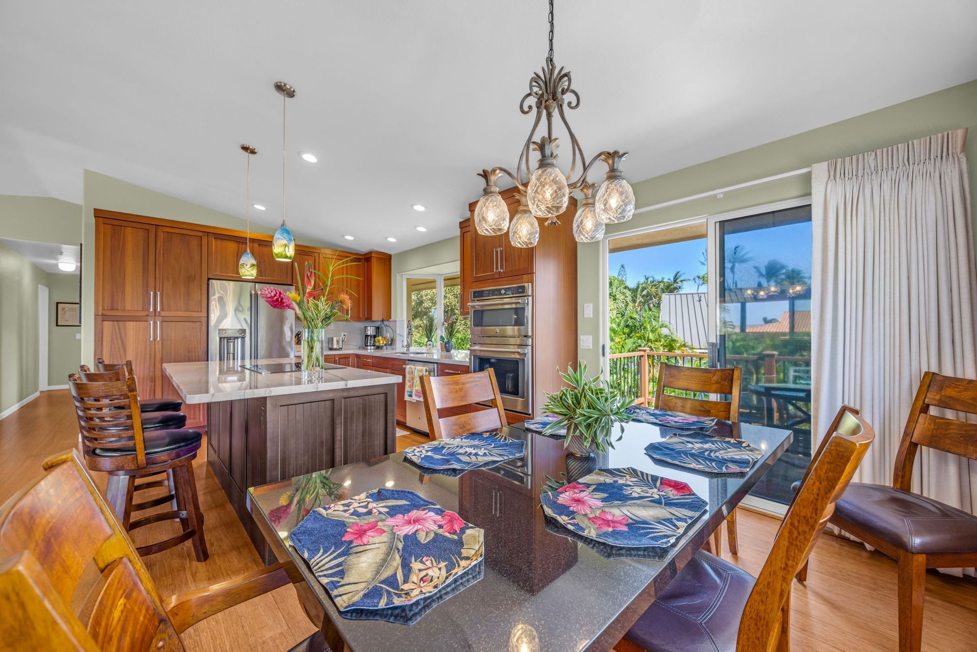 A dining area with a glass table, wooden chairs, and a chandelier, opening to a kitchen with wooden cabinets and island.