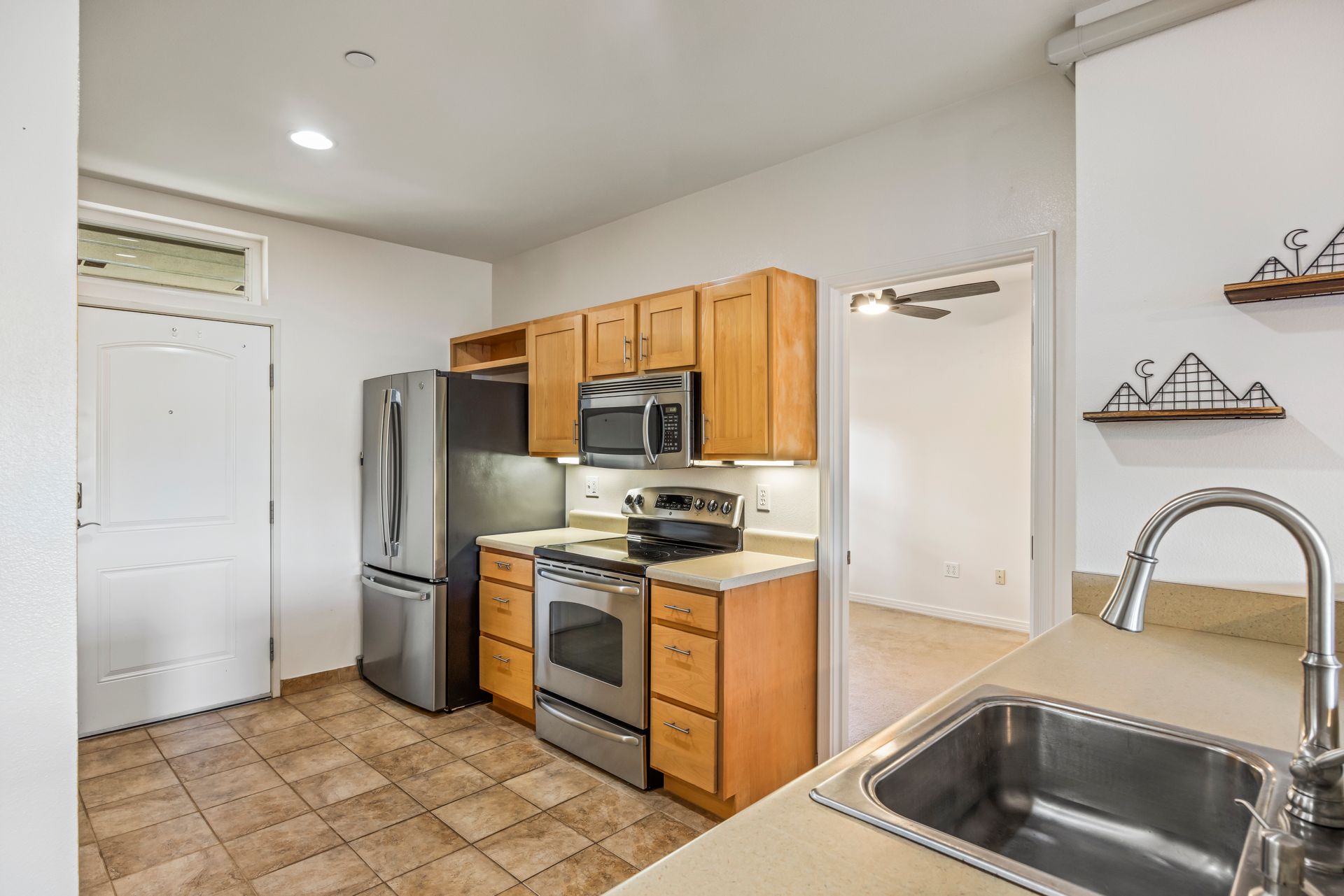 A kitchen with light wood cabinets, stainless steel appliances, tile flooring, and a view into an adjacent room.