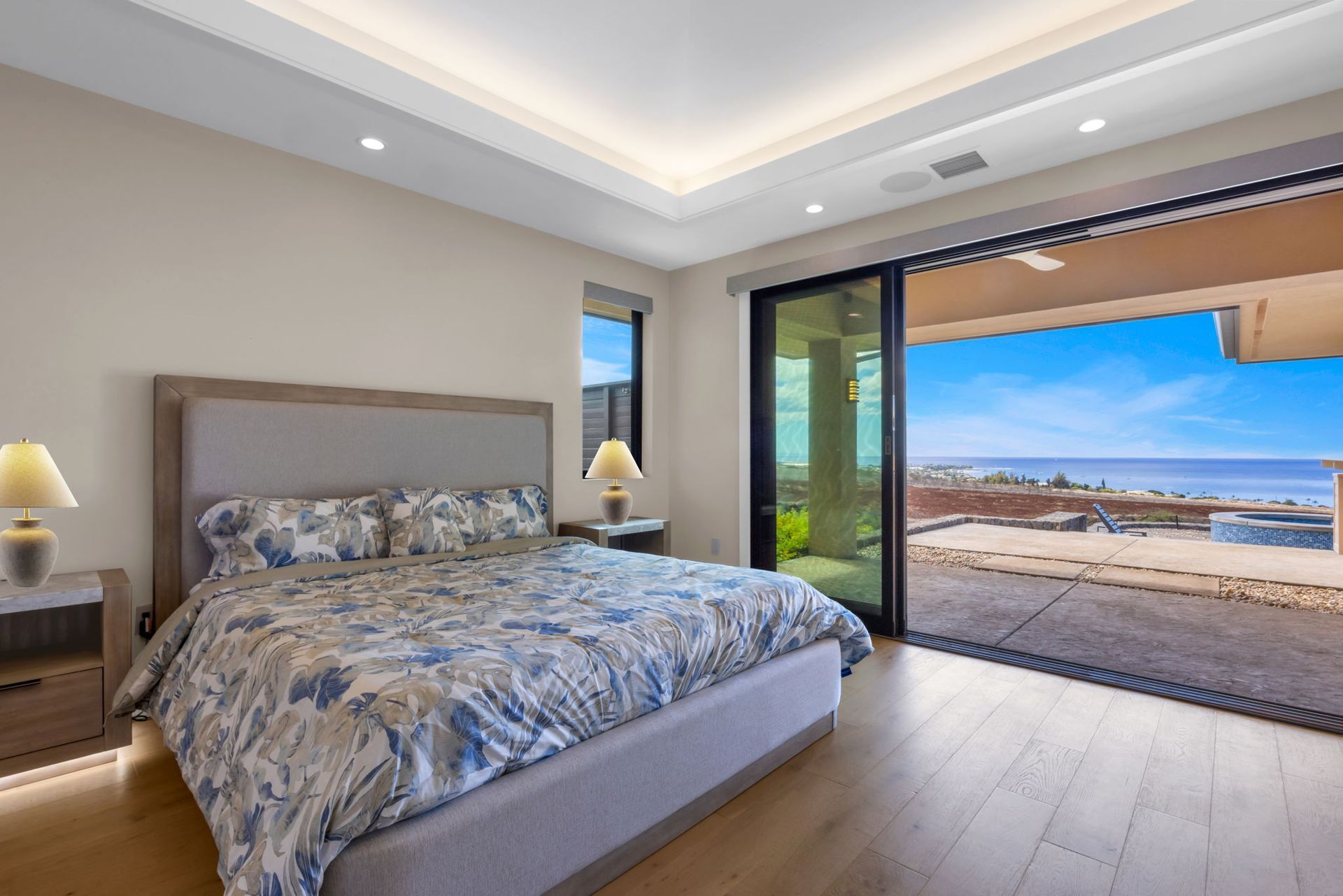 A modern bedroom with a large bed, two lamps, and wide sliding glass doors opening to a coastal view with an ocean horizon.