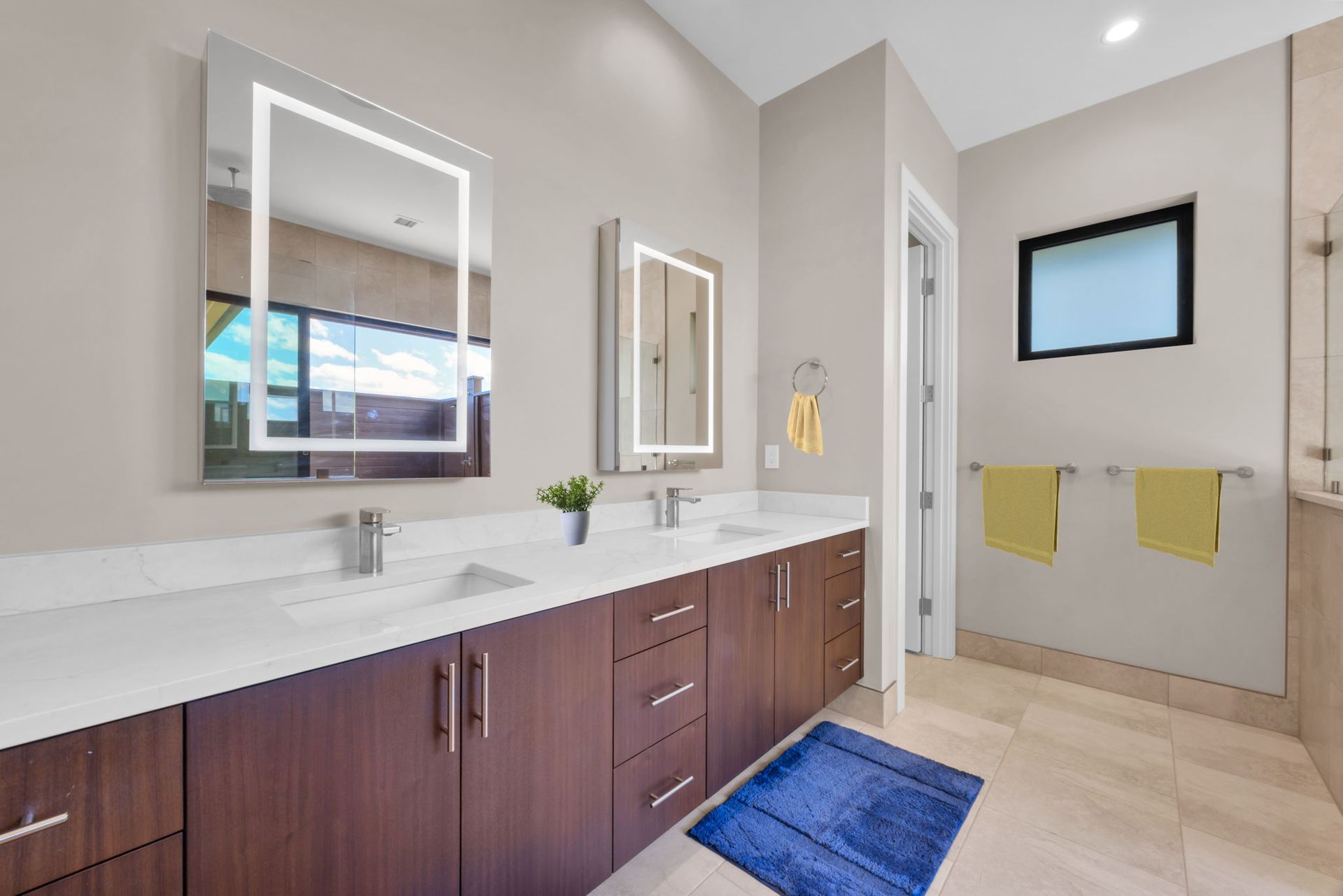 A modern bathroom featuring a double-sink vanity with dark wood cabinets, backlit mirrors, and beige tiled flooring.