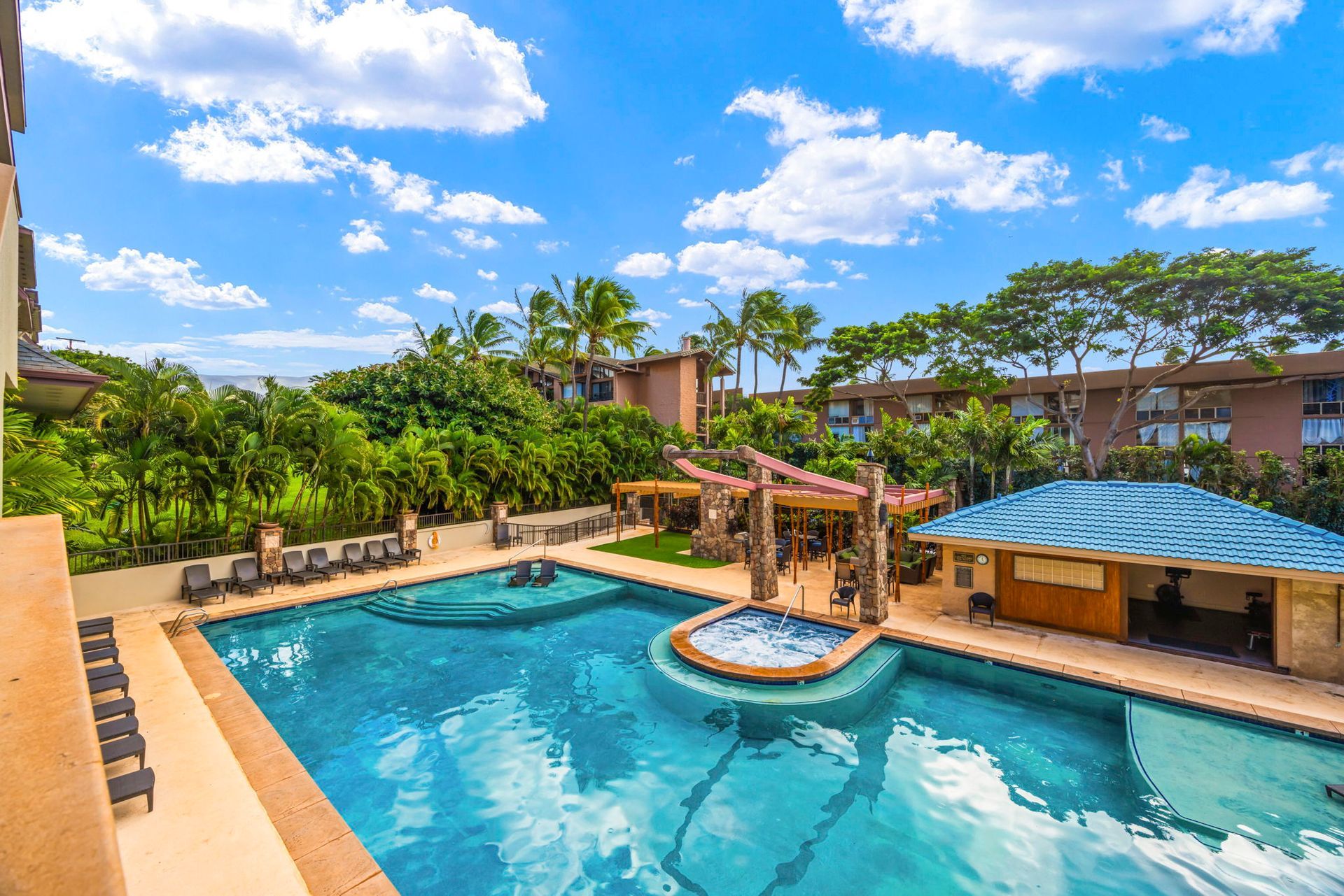A resort swimming pool with a central hot tub, surrounding lounge chairs, and a covered pool bar under a blue sky.