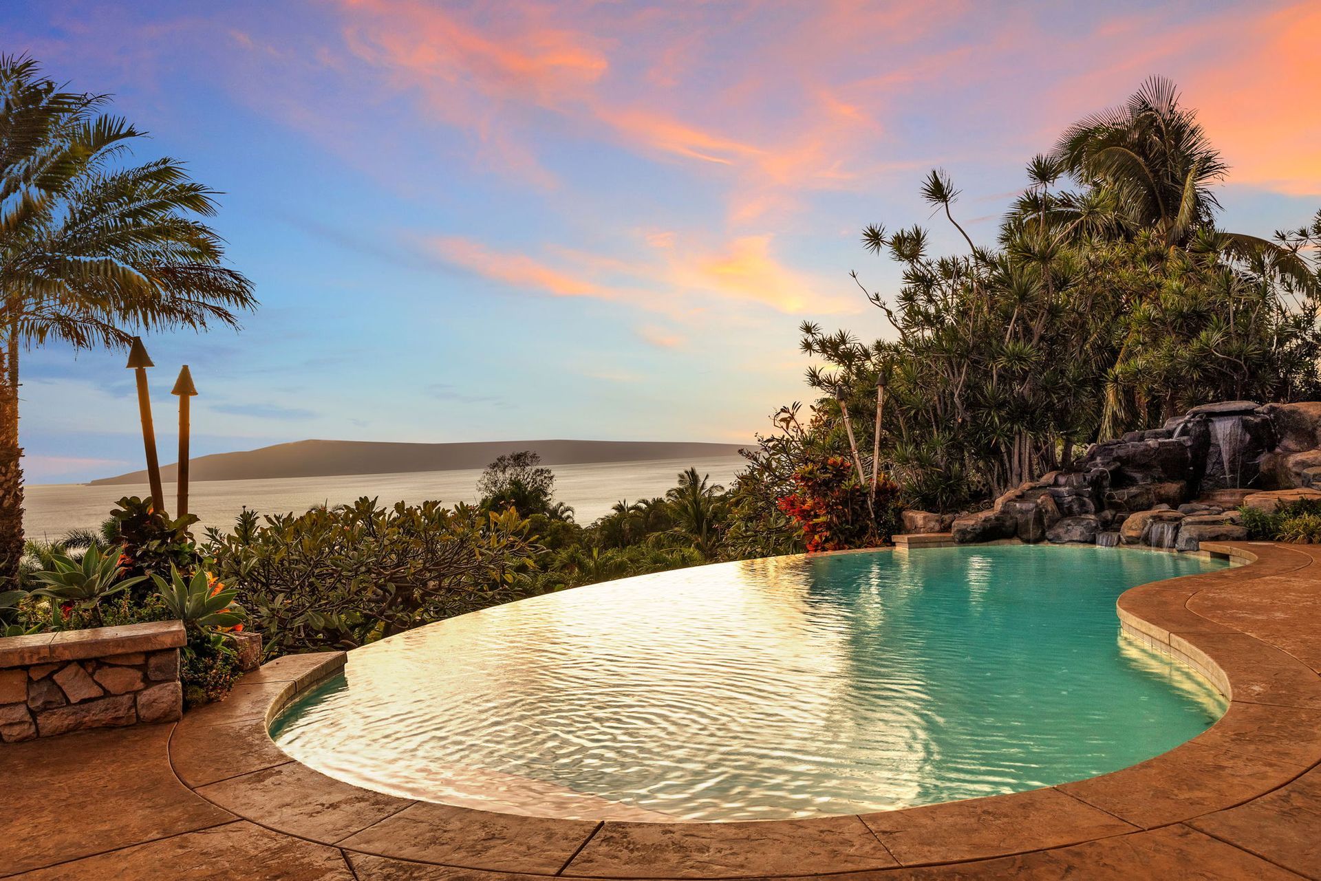 A curved infinity pool overlooks a foggy landscape and distant hills during a colorful orange and blue sunset.
