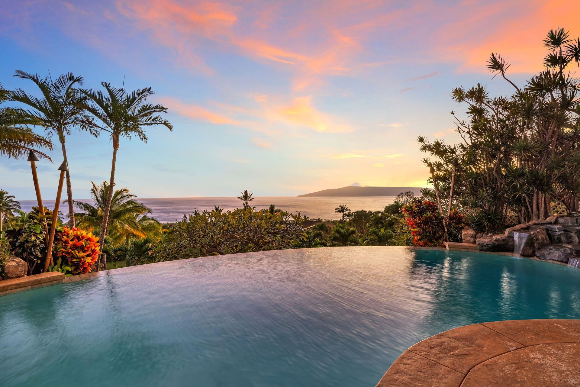 An infinity pool overlooks a lush coastline and ocean during a vibrant sunset with pink and orange clouds.