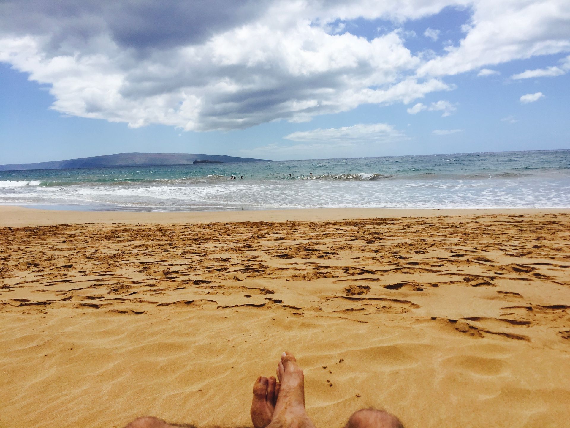 A person's feet resting on a sandy beach, overlooking the ocean under a cloudy sky.