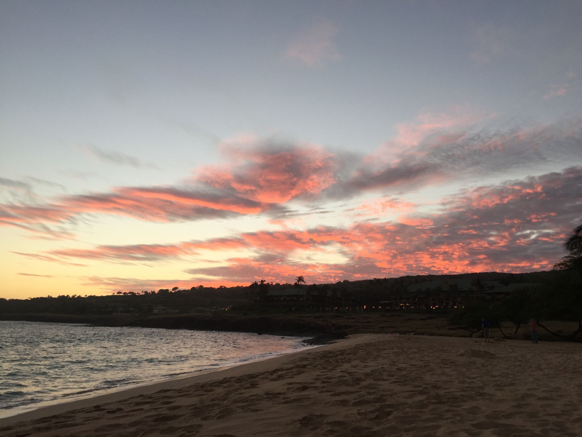 A sandy beach at sunset with pink and orange clouds streaking across the sky above a dark shoreline.