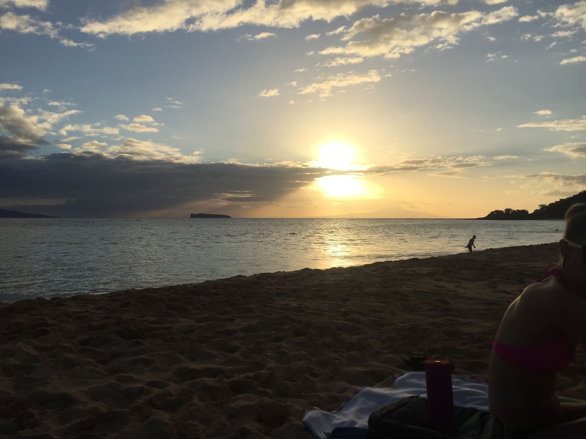 A scenic beach at sunset with a person in the foreground, calm ocean water, and soft clouds in the sky.