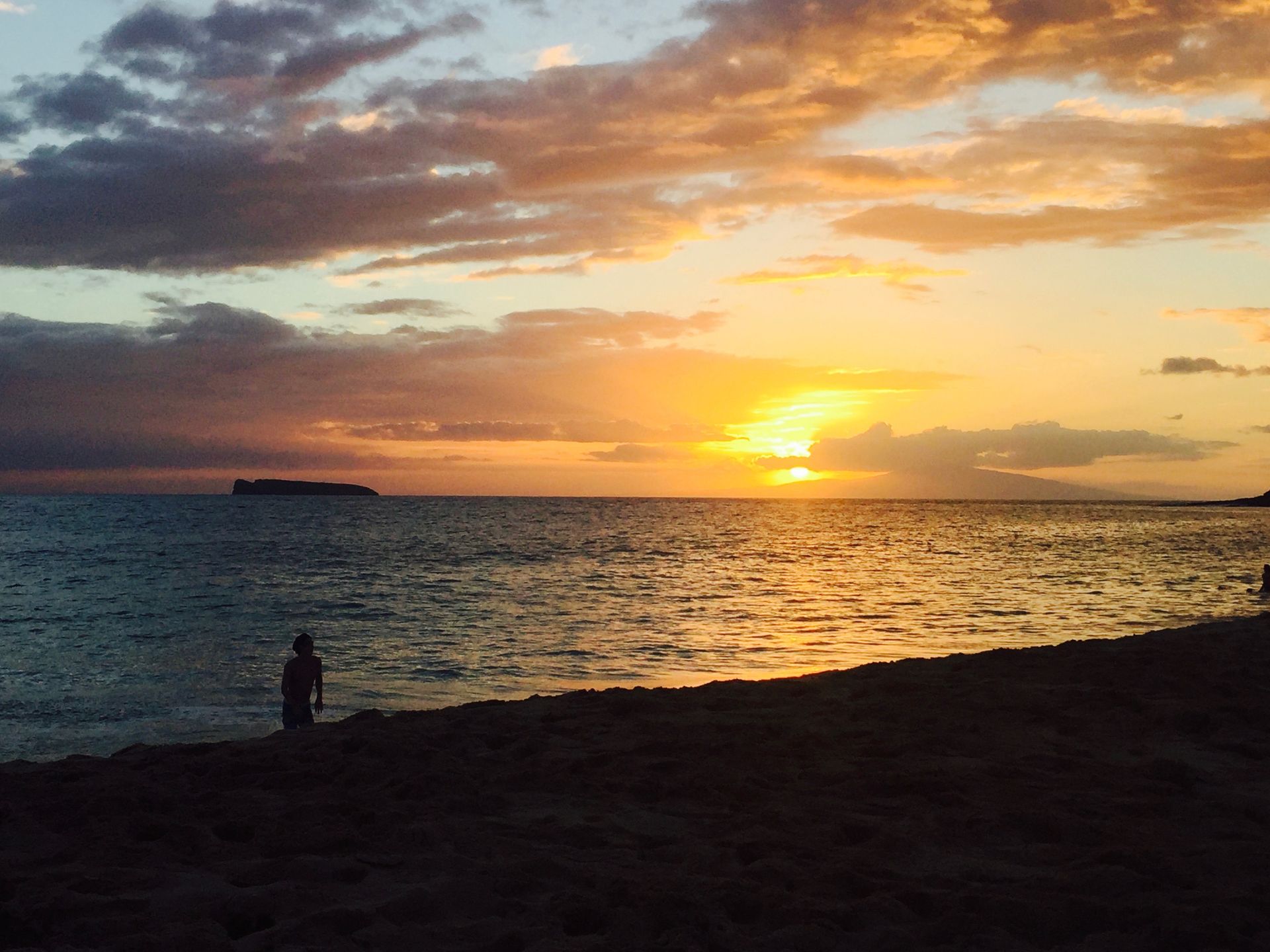 A person stands on a sandy beach at sunset, looking out at the ocean with distant islands on the horizon.