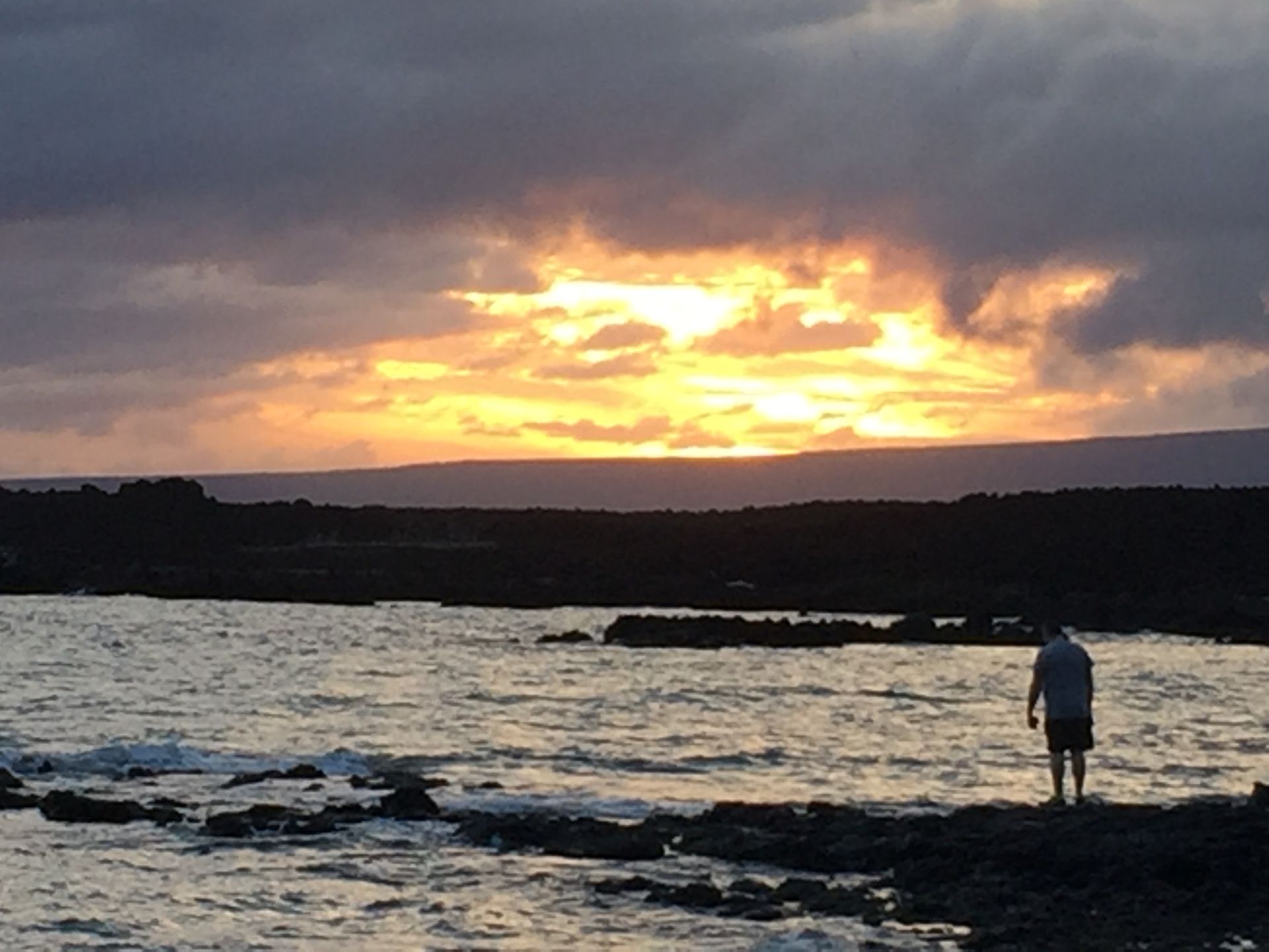 A person stands on a rocky shore watching a golden sunset over the ocean beneath a cloudy sky.