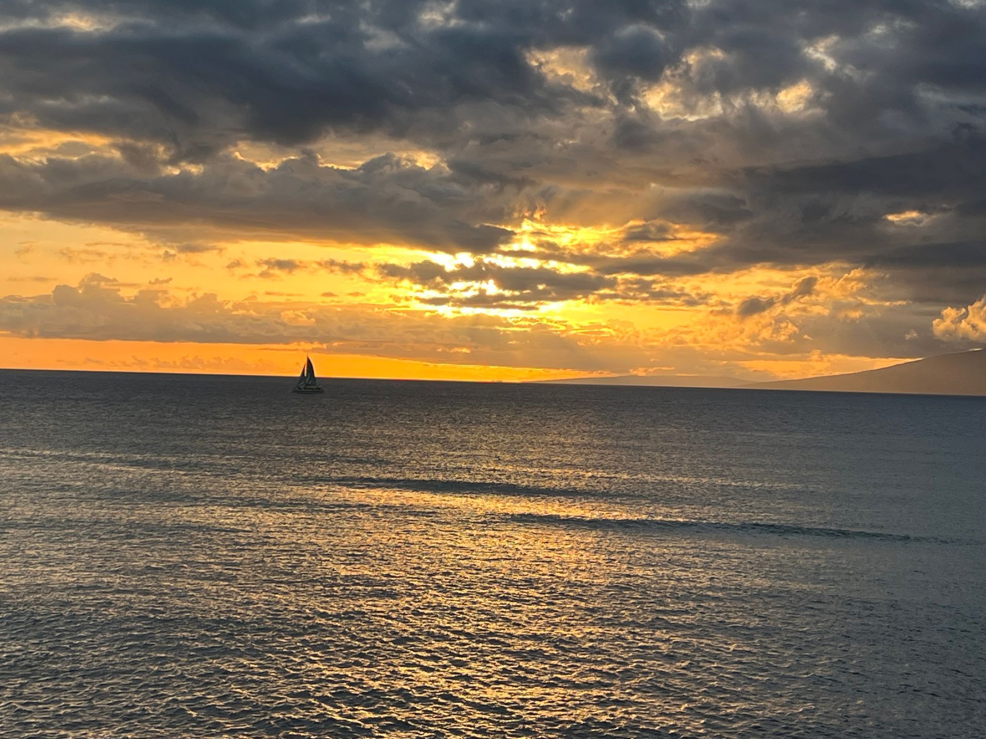 A sailboat drifts on the ocean during a vibrant golden sunset beneath a sky filled with dark, dramatic clouds.