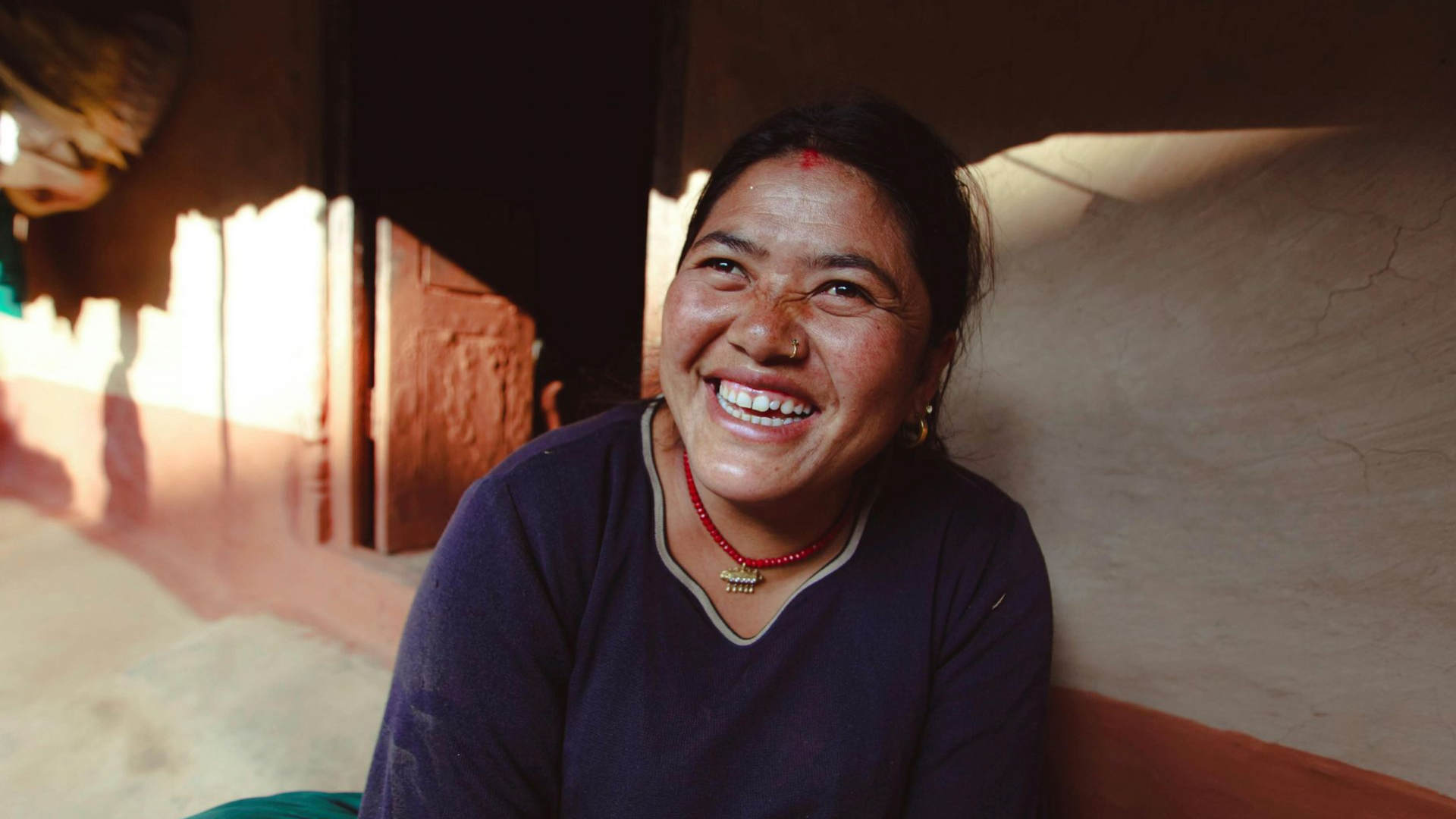 A person with a wide, joyful smile, wearing a dark top and a red beaded necklace, sits indoors against a textured wall.