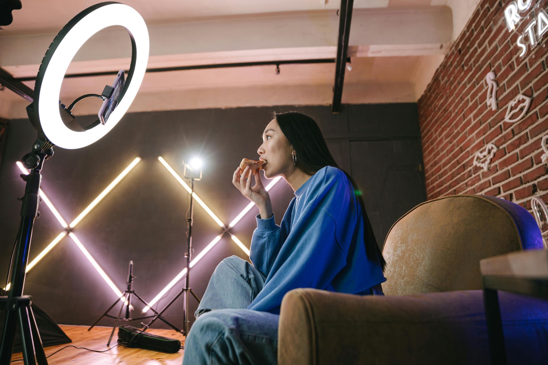 A person in a blue sweater sits in a studio with ring lighting, eating a snack while facing a camera.