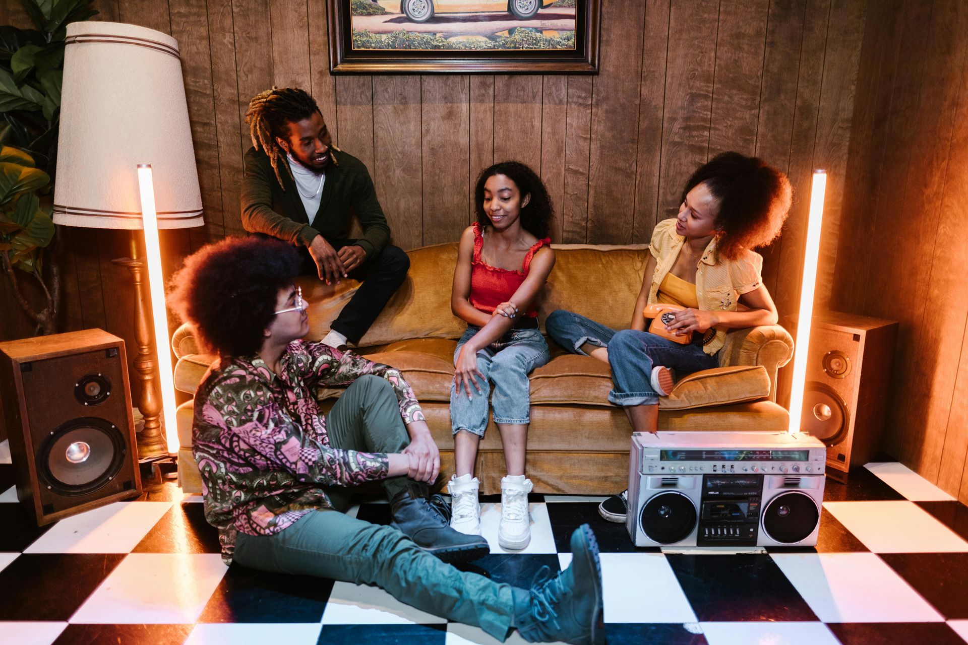 Four people sit in a room with a checkerboard floor, vintage speakers, and warm lighting, conversing on and near a sofa.