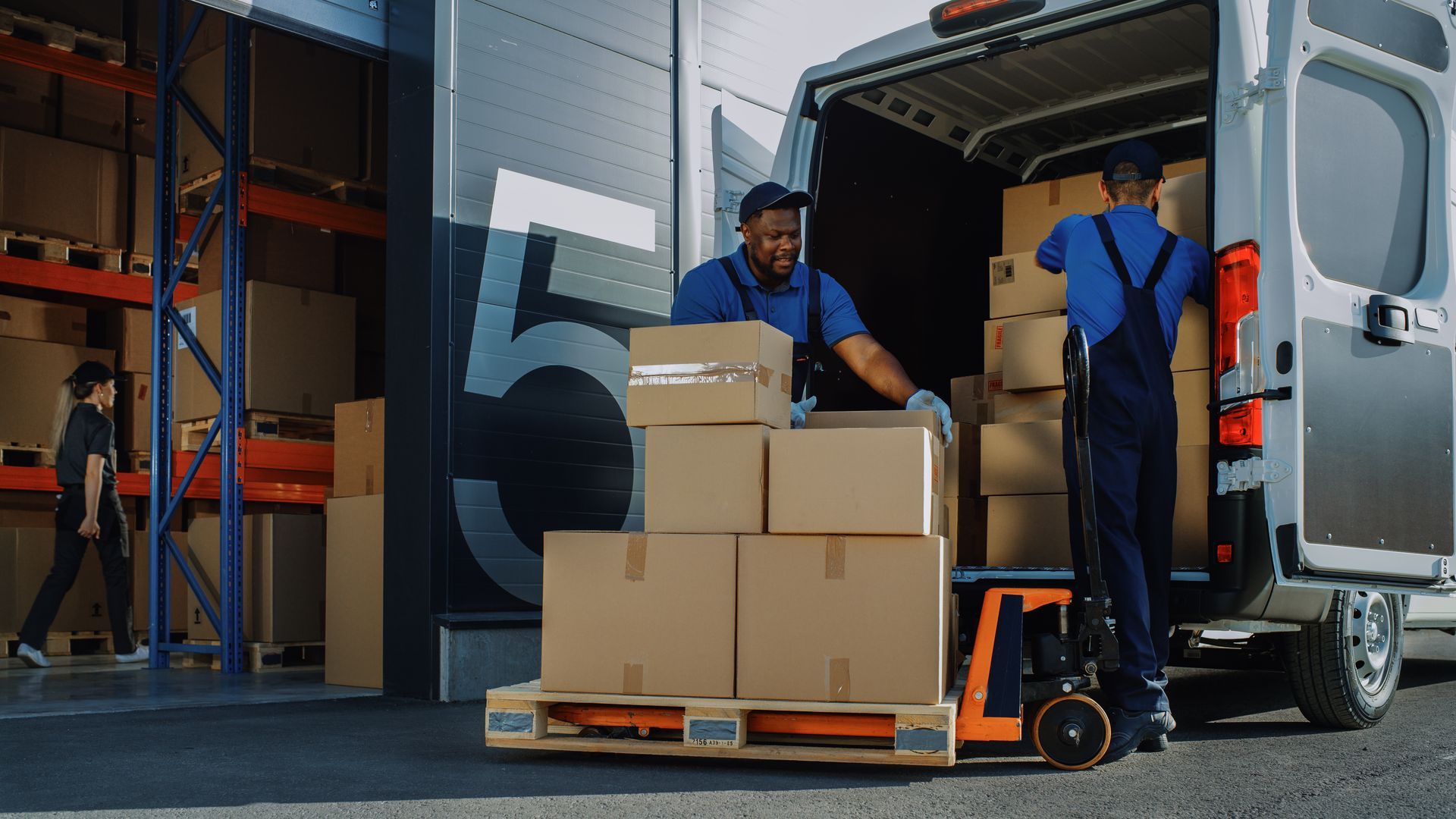 Workers loading boxes onto a pallet jack beside a delivery truck at a warehouse dock.