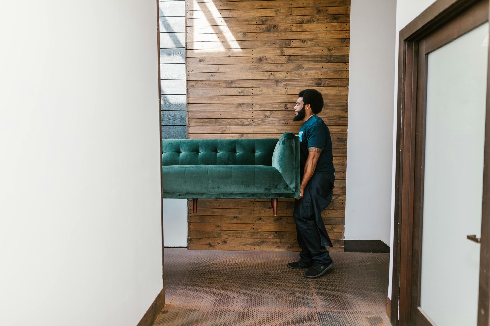 Person carrying a teal sofa through a hallway with wood-paneled wall