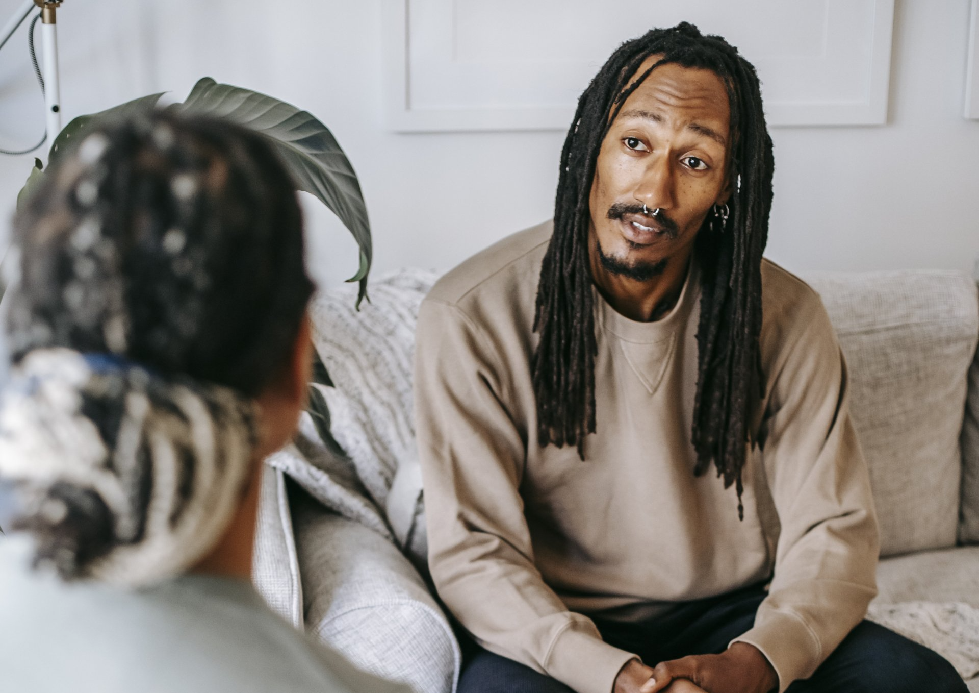 A man with dreadlocks is sitting on a couch talking to a woman.