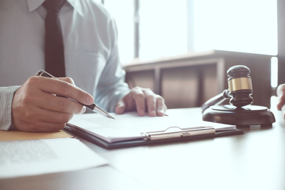 A Man is Sitting at a Table Writing on a Clipboard Next to a Judge's Gavel — ACT Law Associates In Canberra, ACT