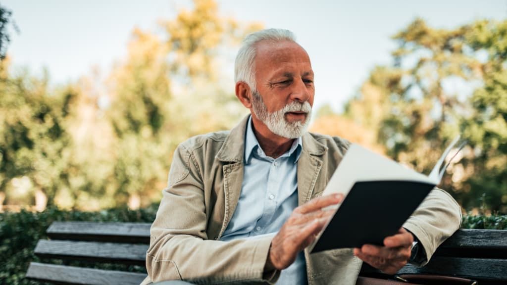 An Elderly Man Is Sitting On A Park Bench Reading A Book— ACT Law Associates In Canberra, ACT