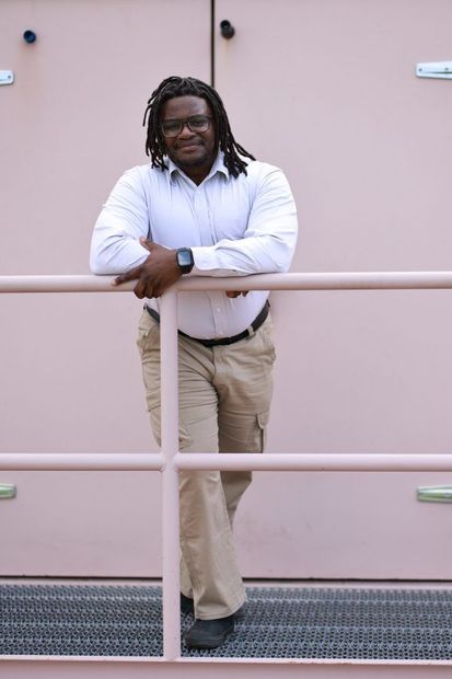 Man in a light blue shirt and khaki pants leans on a pink railing, smiling at the camera.