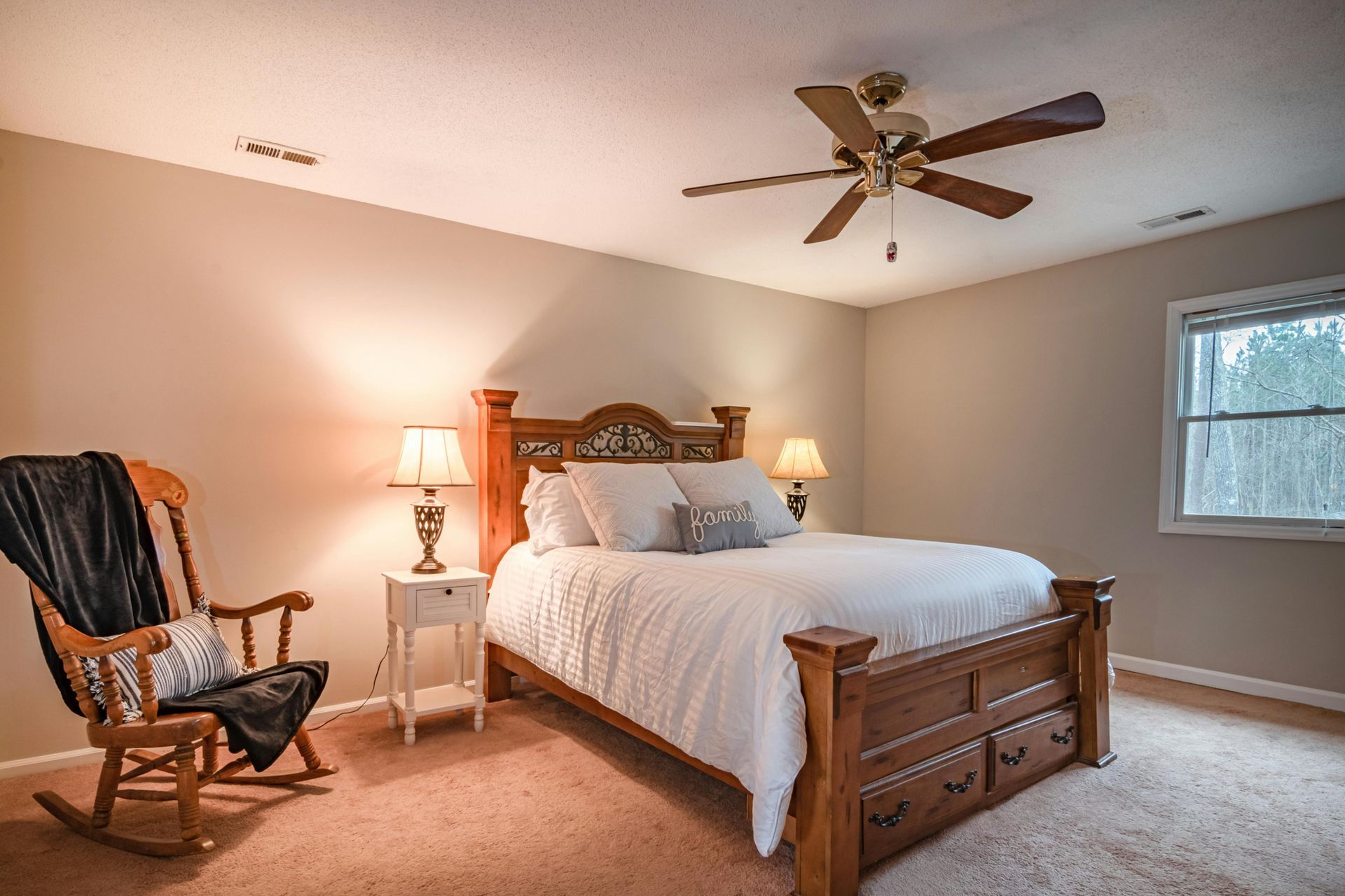 Bedroom with wooden bed, nightstand, rocking chair, and ceiling fan. Light brown walls and carpet.