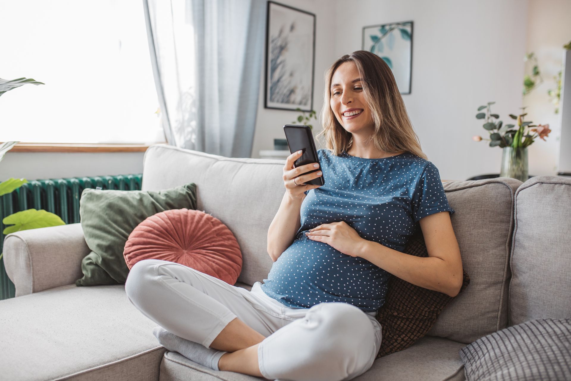 A pregnant woman is sitting on a couch looking at her cell phone.