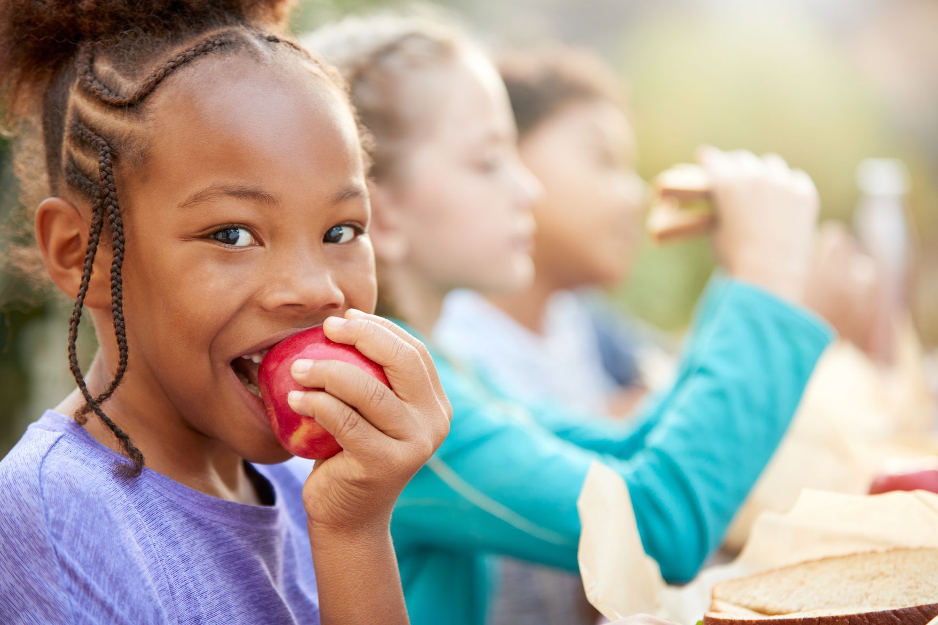 Girl sitting at a lunch table with other children and biting into a red apple