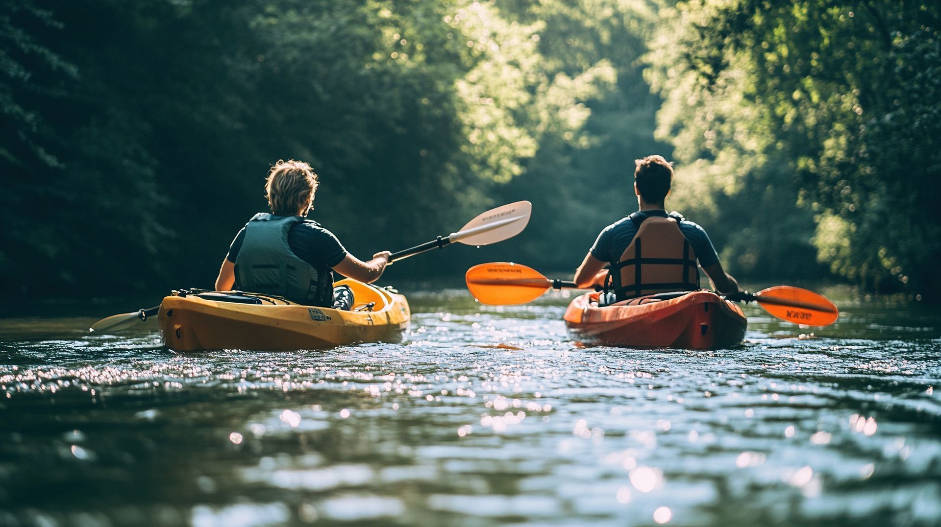 Two kayakers paddling on a sunlit river, seen from behind, with a forested shoreline ahead.