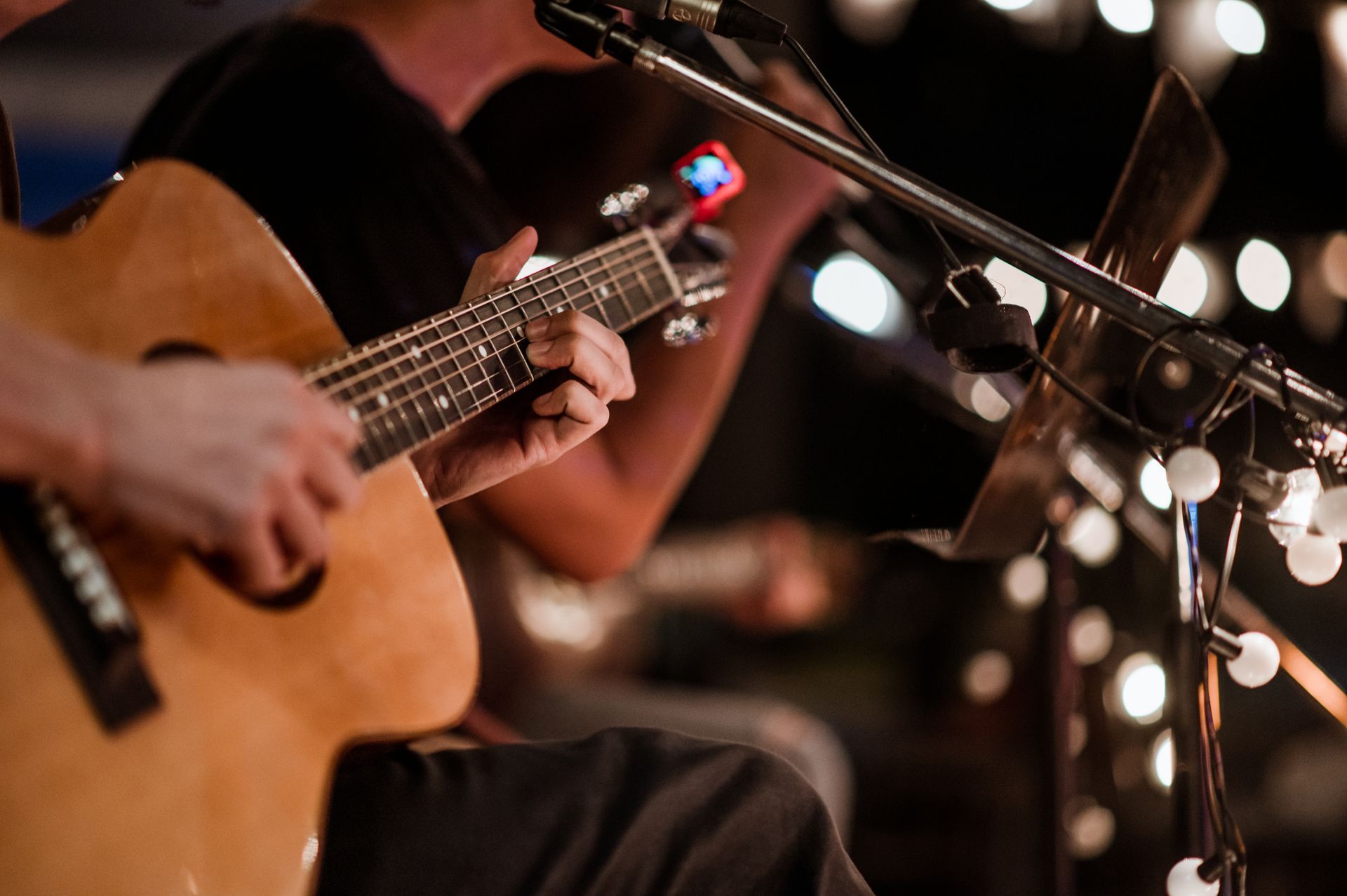 Musician strumming an acoustic guitar on stage with blurred lights and a microphone stand nearby