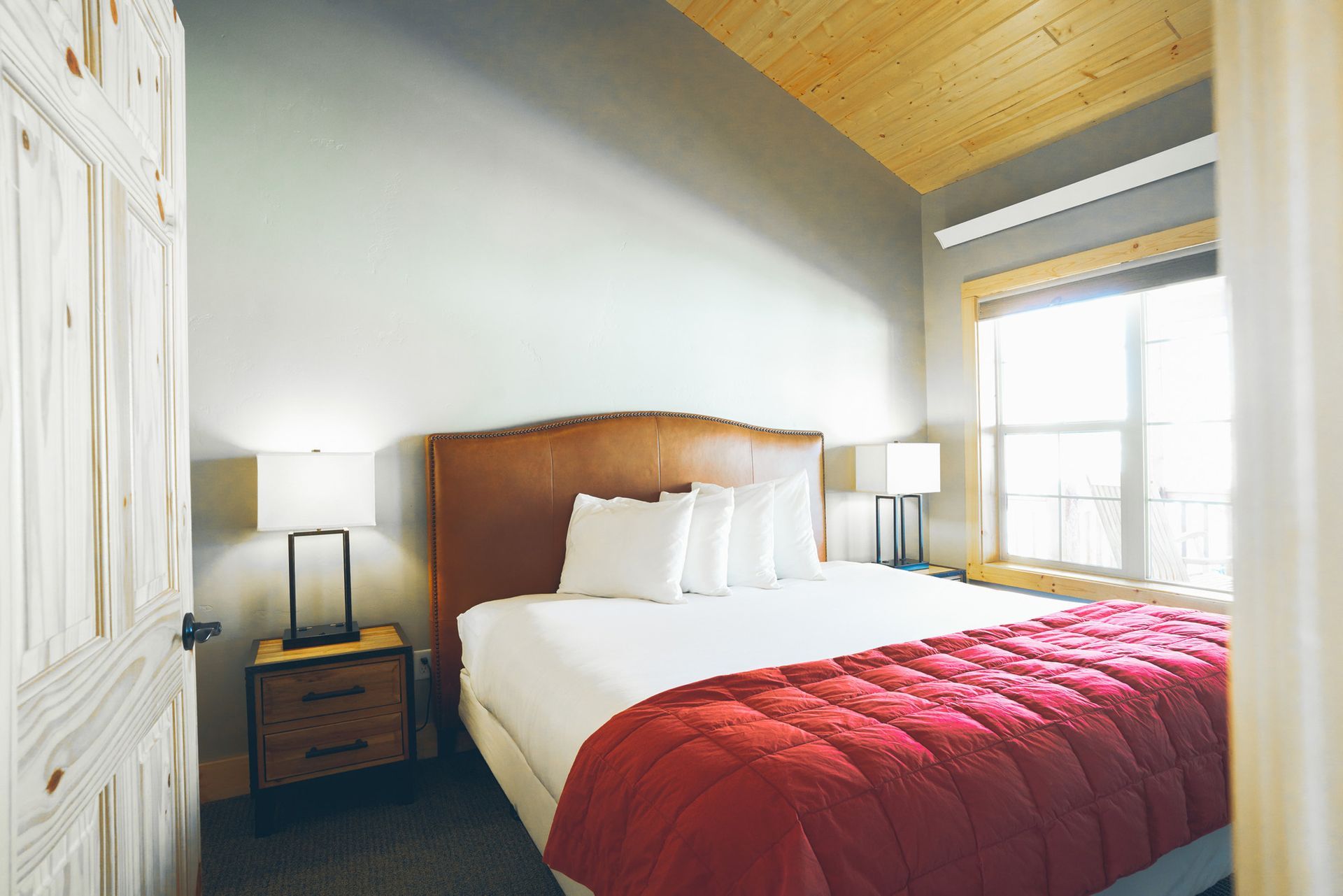 Cozy bedroom with a wooden bed, red quilt, bedside lamps, and bright window light