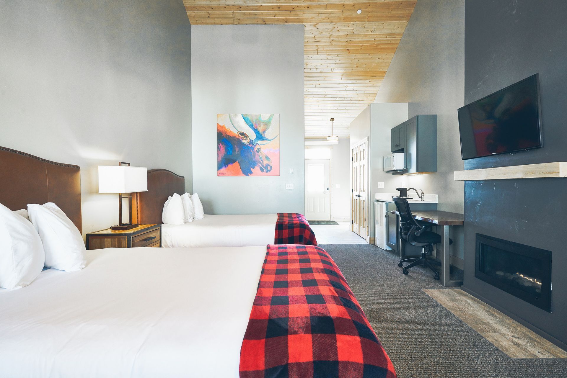 Modern hotel room with two beds, TV, fireplace, and red plaid runner, viewed toward a bright doorway