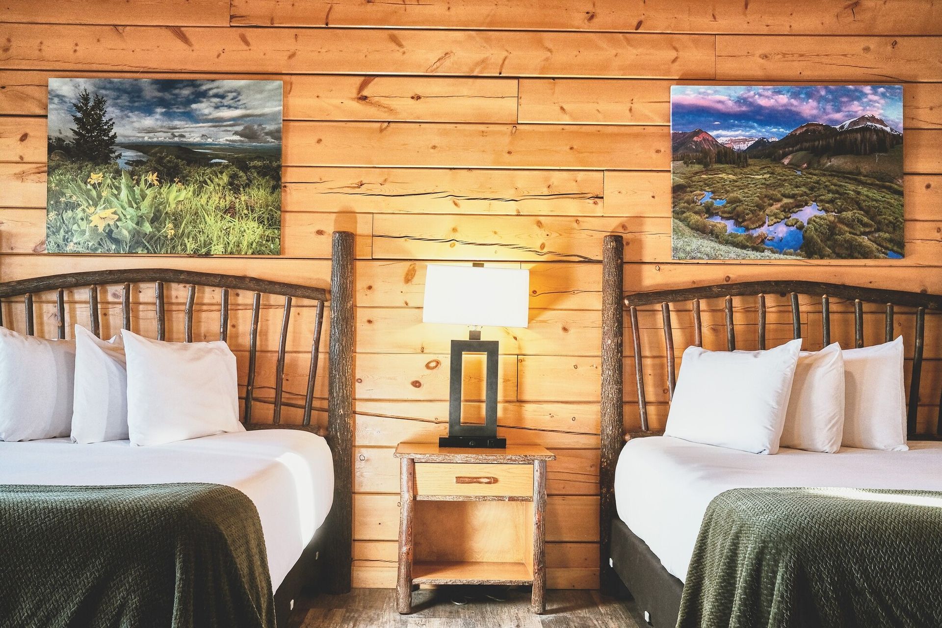 Cozy cabin bedroom with two twin beds, wood-paneled walls, lamp, and landscape paintings