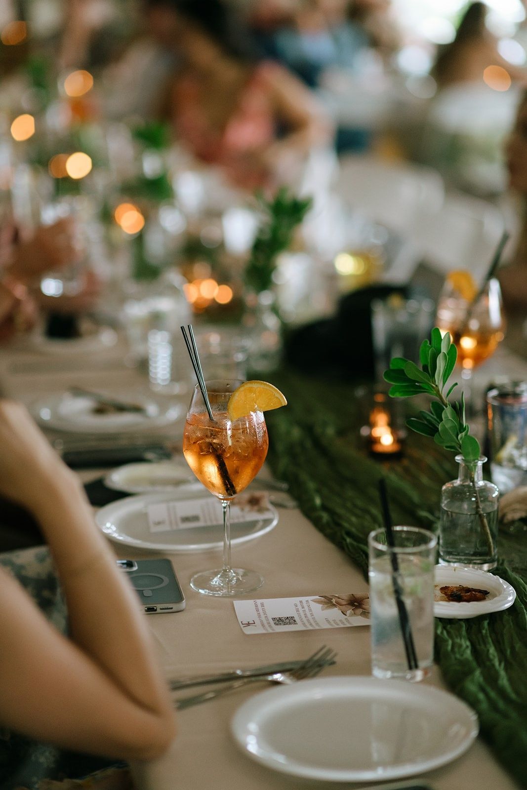 Elegant candlelit dinner table with white plates, green floral runner, and orange cocktails in coupe glasses