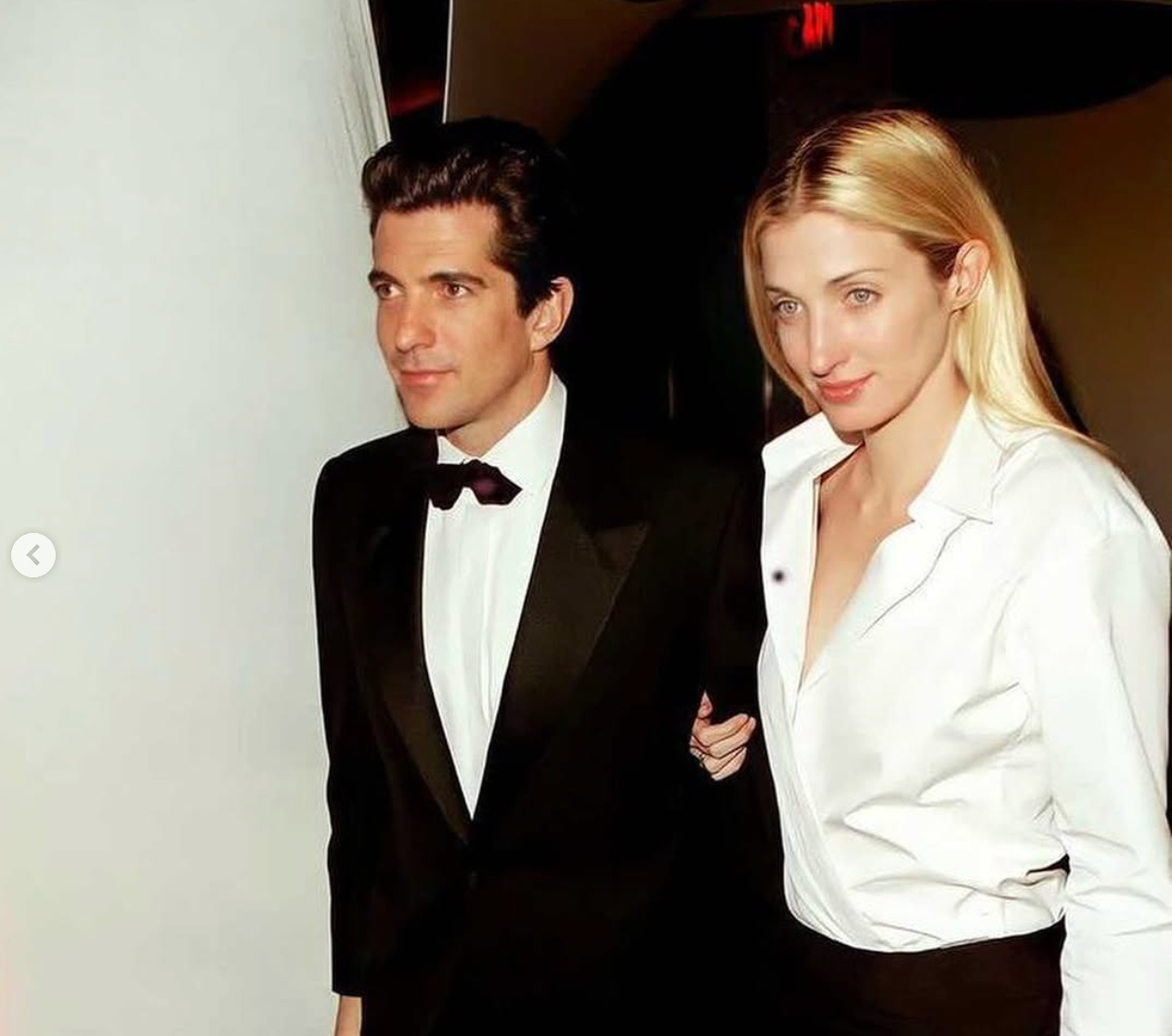 JFK Jr. wearing a tuxedo walking with Carolyn Bessette wearing a white blouse and black skirt.