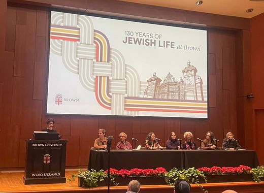 Leora Tanenbaum standing at podium with 7 panelists seated to her left at the Brown University event in November 2025, 