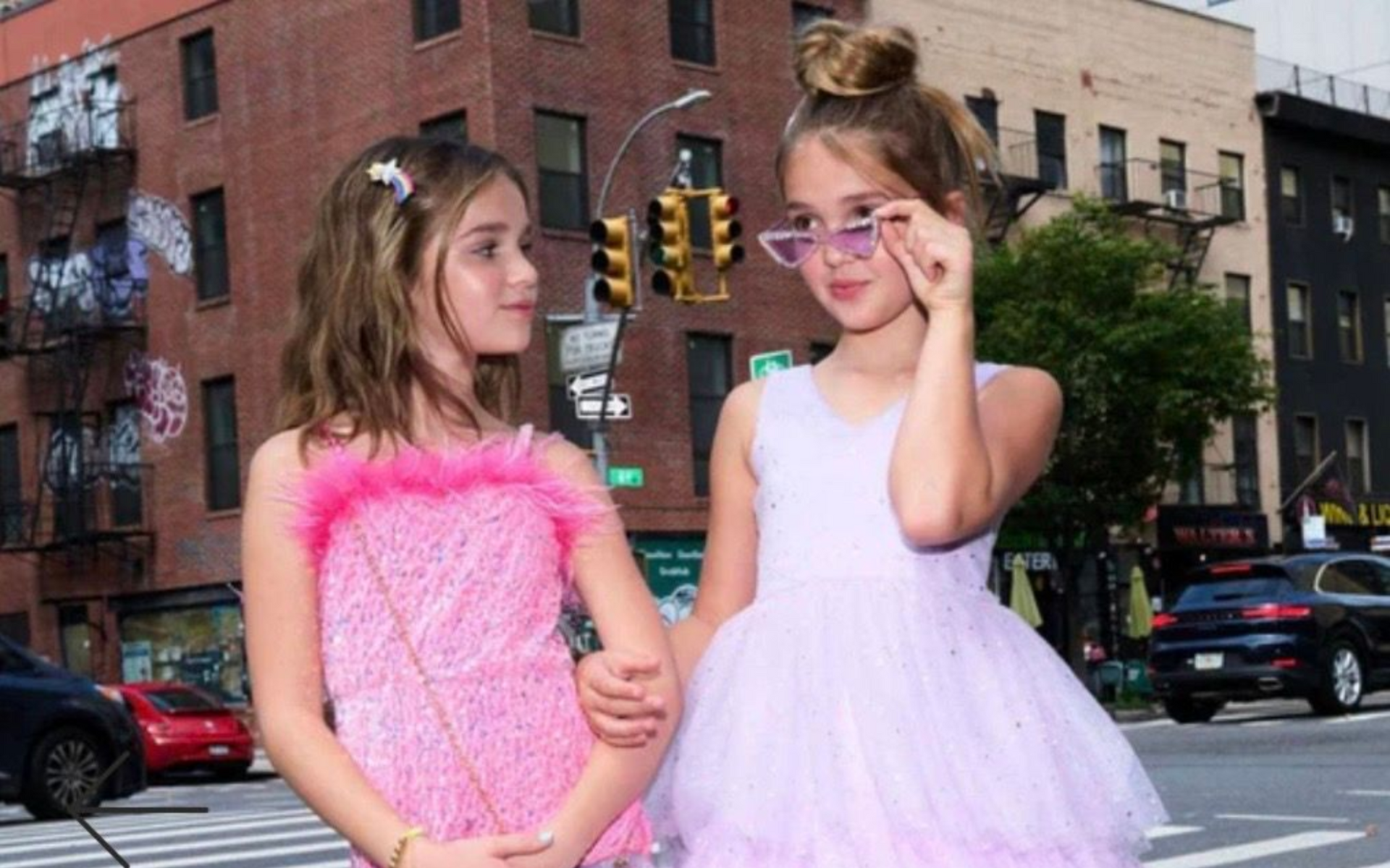 Two white girls, approximately age 10, in pink party dresses standing outside in urban landscape.