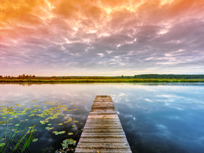 Wooden dock extends into a calm lake, reflecting a colorful sunset sky.